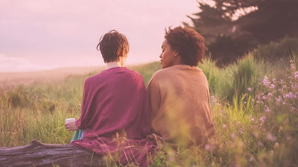 Two adults in relationship sit together on a driftwood log by the sea, surrounded by sea grass and trees | Blog | CSP