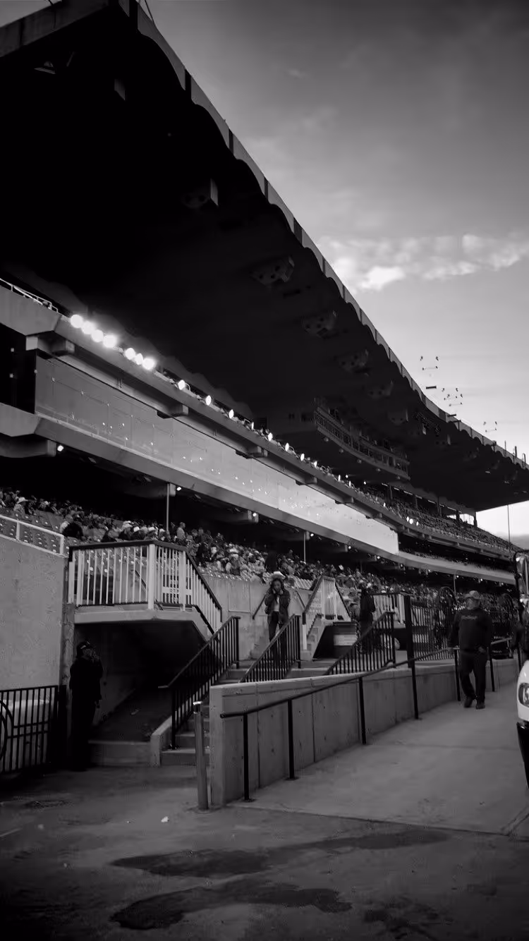 Black and white photo of a stadium's covered seating area with spectators and people walking near staircases.