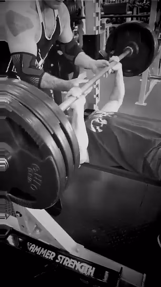 Man lying on a bench performing a heavy barbell bench press with spotter assisting in a gym.