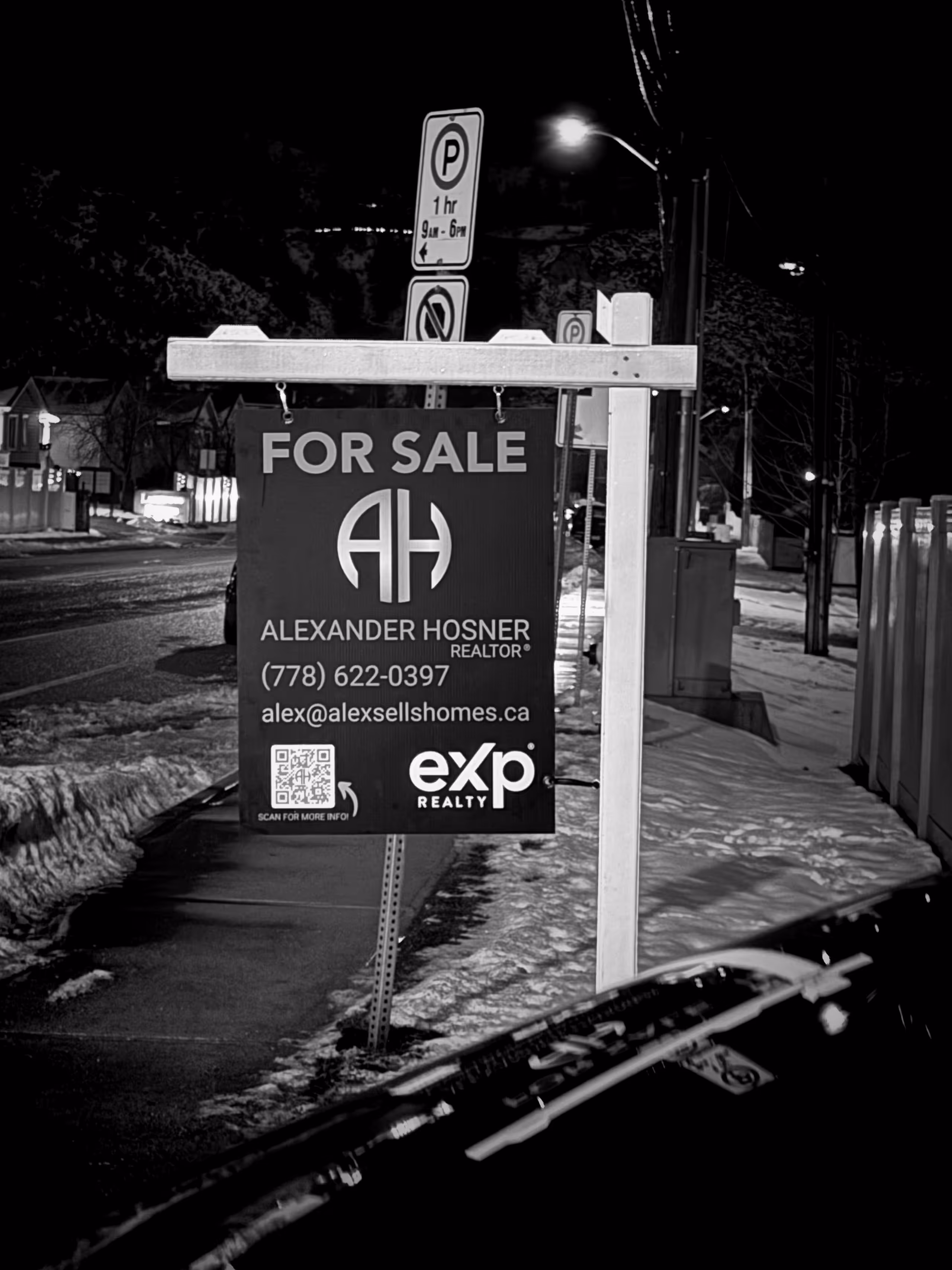 Real estate for sale sign for Alexander Hosner with contact info, hanging on a post by a snowy sidewalk at night.