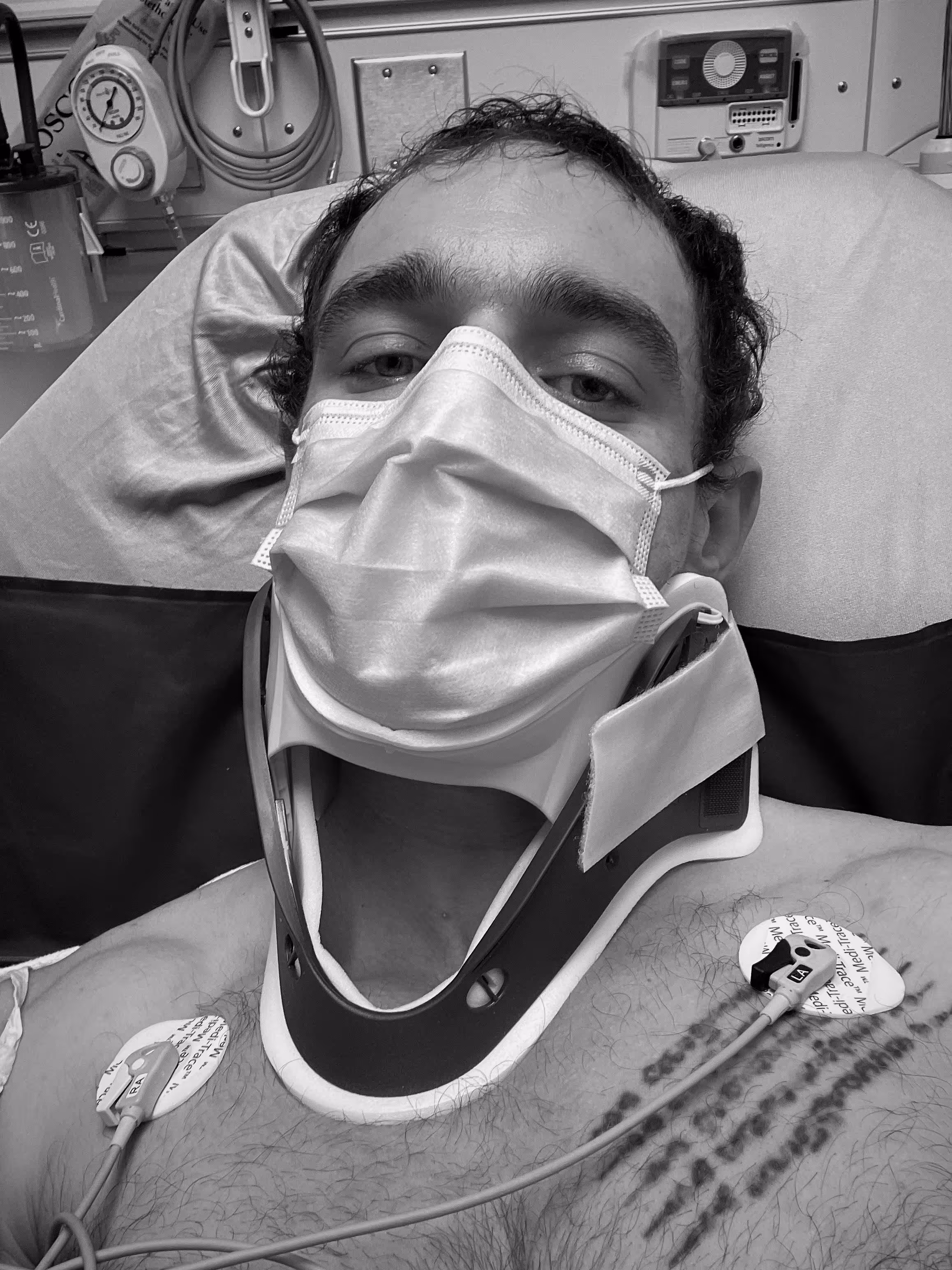 Man wearing a medical face mask and a neck brace lying in a hospital bed with ECG electrodes attached to his chest.