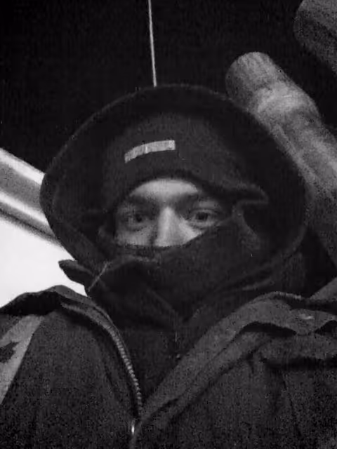 Person wearing a hooded jacket and beanie, with a scarf covering the lower half of their face, in a low-light setting.