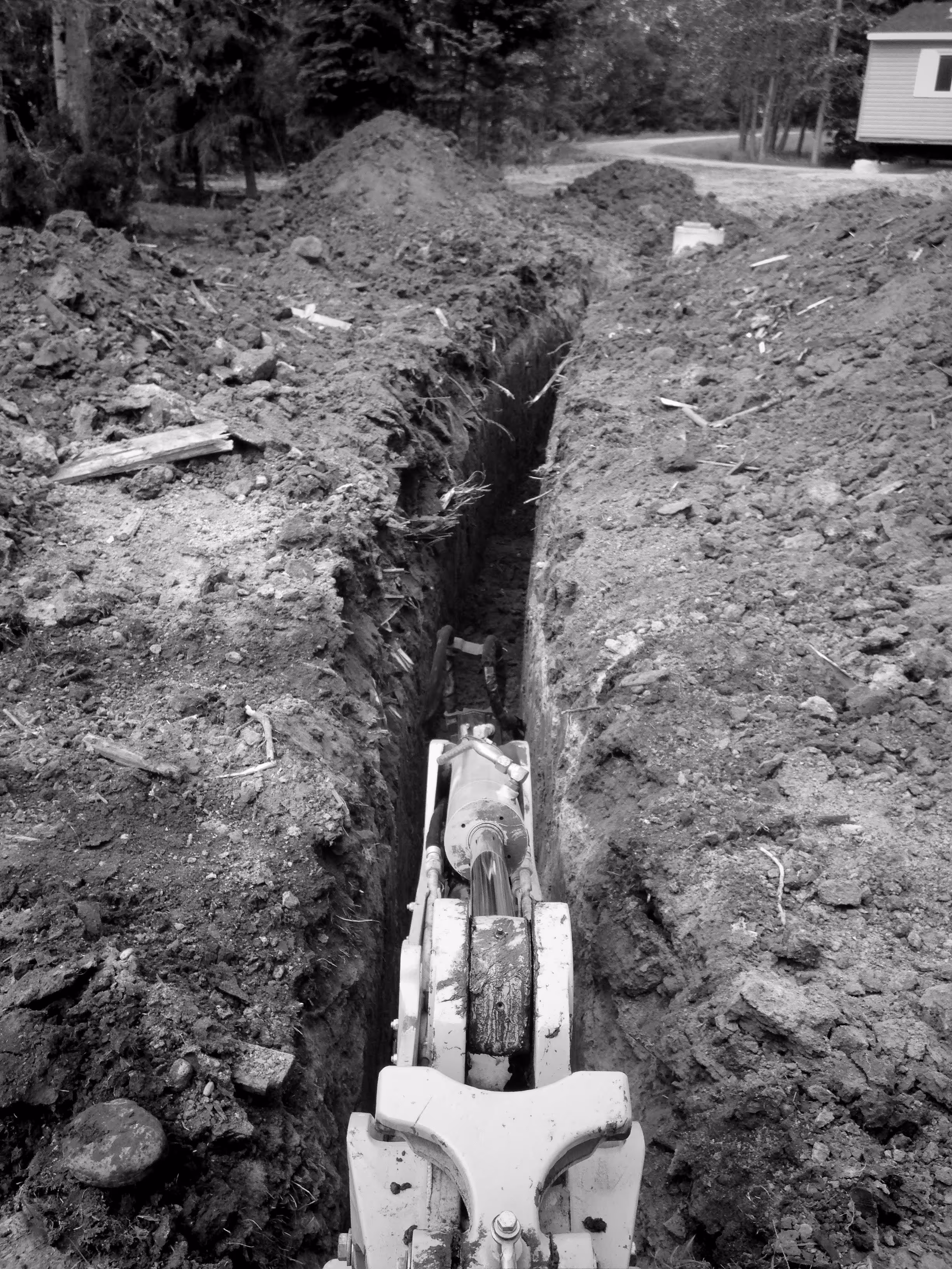 Close-up of an excavator arm digging a narrow trench in soil near a wooded area with a house in the background.