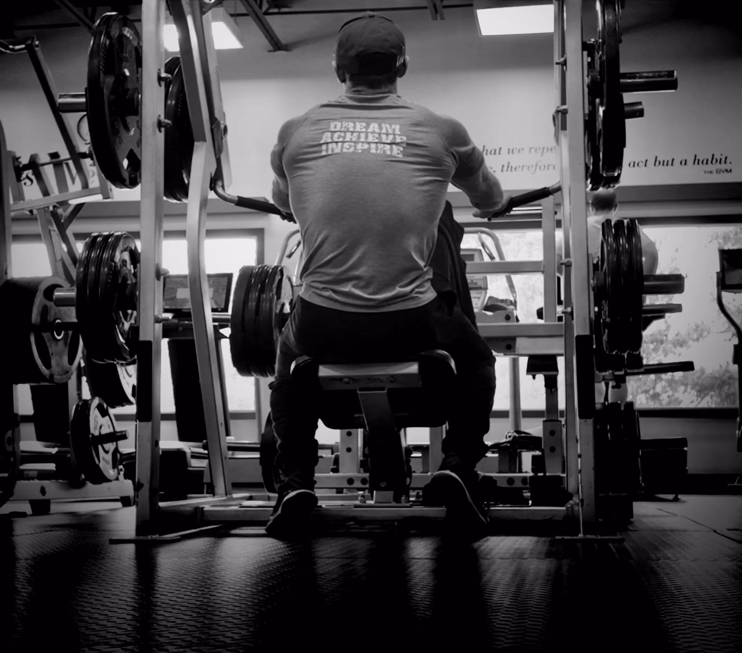Man wearing a cap and a shirt with the words 'Dream Achieve Inspire' exercising on a seated rowing machine in a gym.
