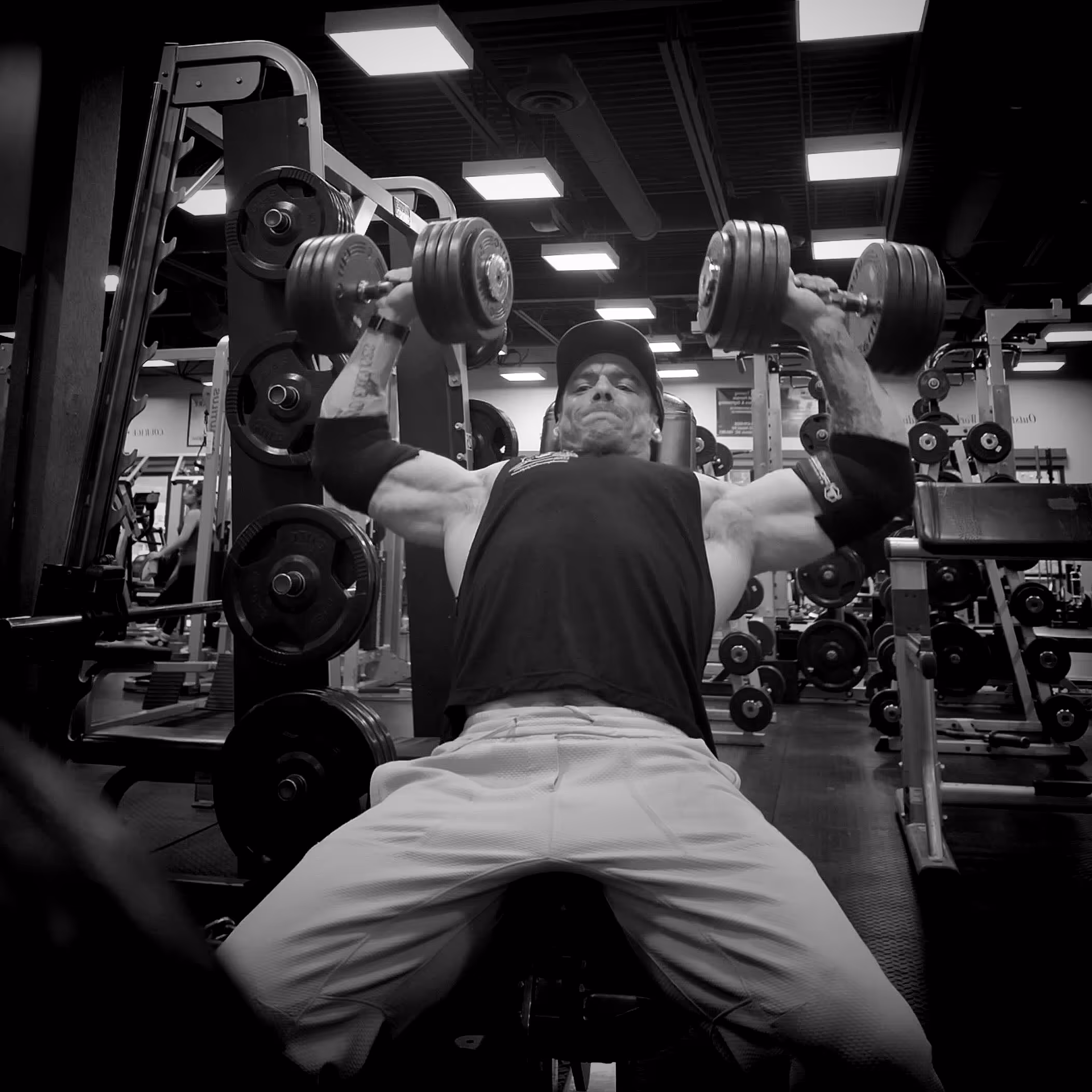 Man in a gym performing an inclined dumbbell press with heavy weights while wearing a cap and elbow supports.