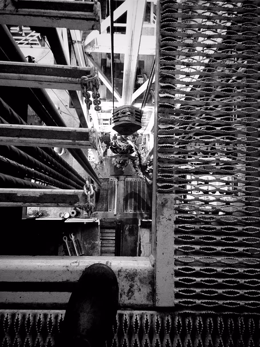 View looking down through a metal grated platform in an industrial setting with safety chains and a booted foot visible.