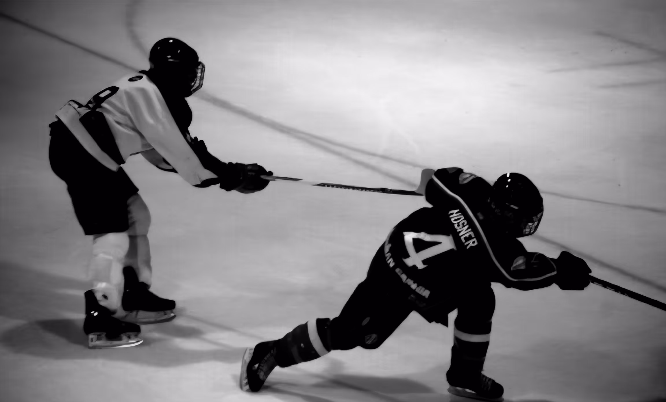 Two ice hockey players compete with one player extending his stick to reach the other player skating forward.