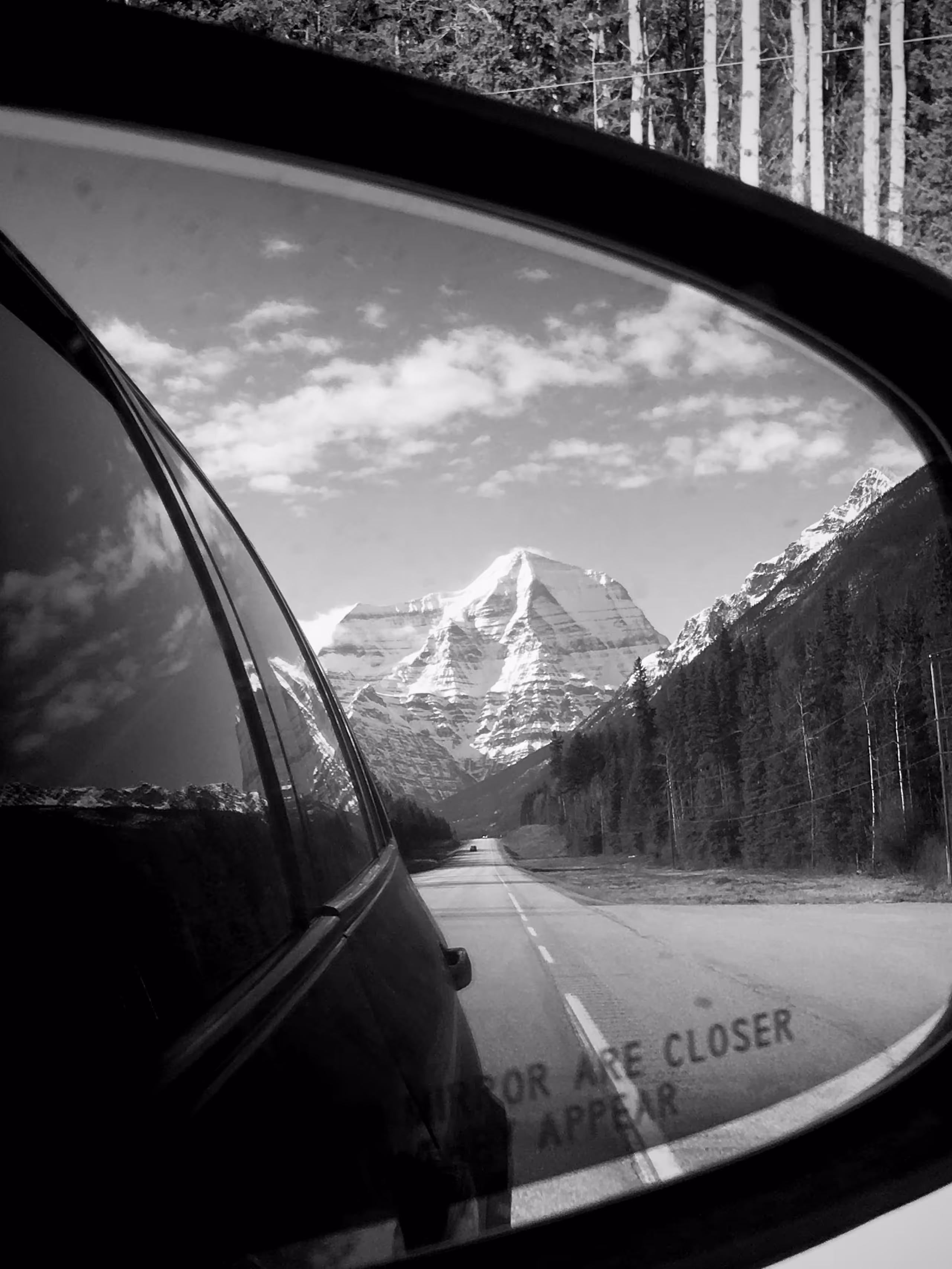 Side mirror reflection showing a car on a road with snow-covered mountains and trees in the background under a partly cloudy sky.