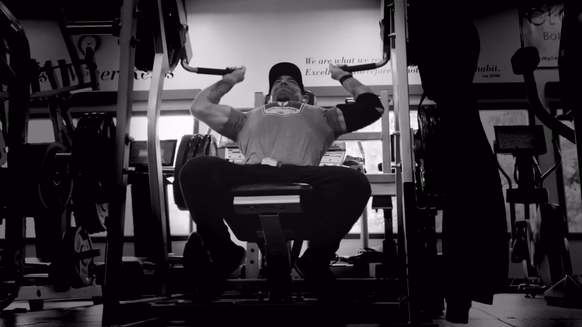 Man wearing a cap and elbow support performing a seated lat pulldown exercise in a gym.
