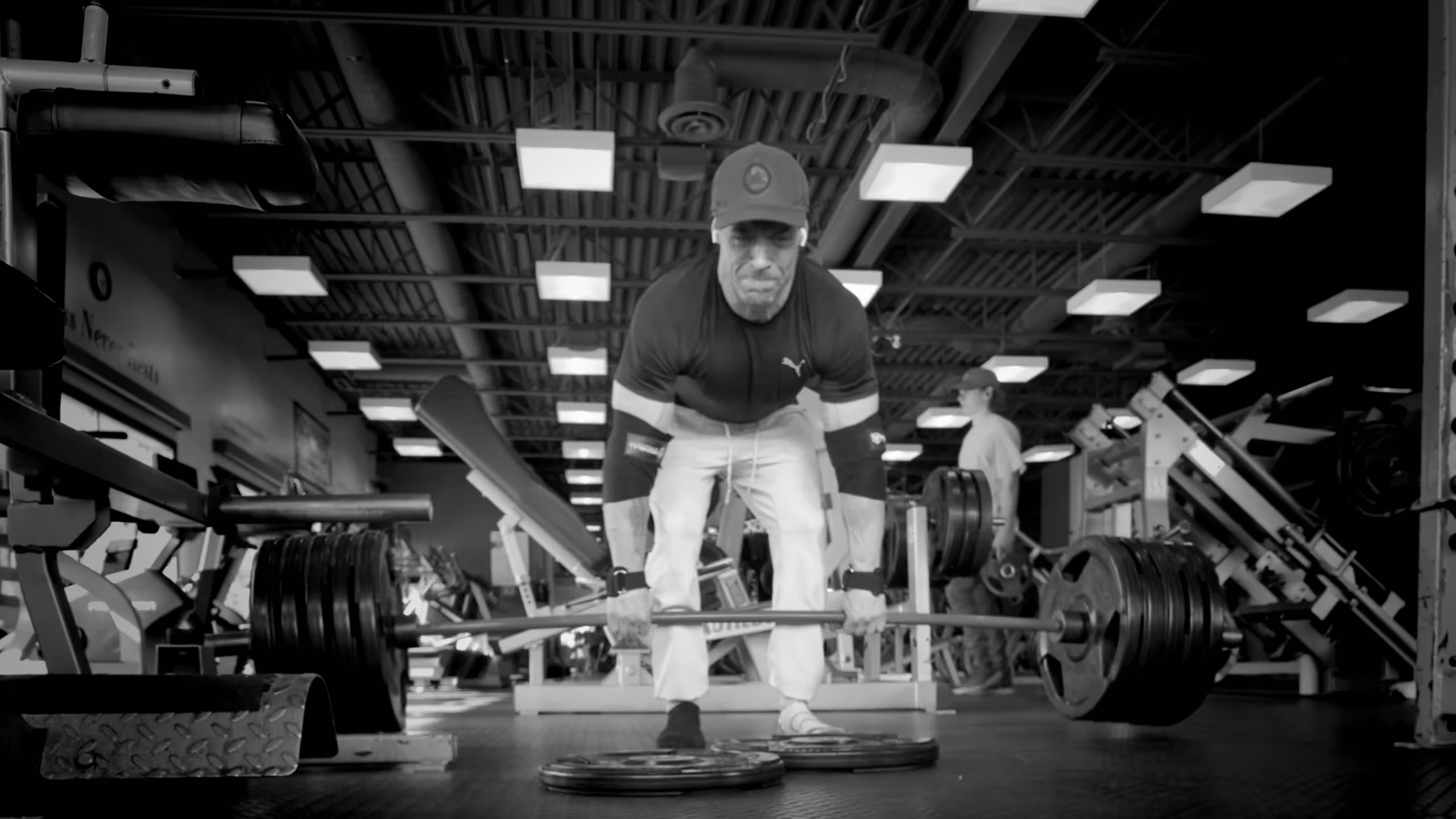 Man in gym lifting a heavily loaded barbell from the floor, wearing a cap and elbow supports.