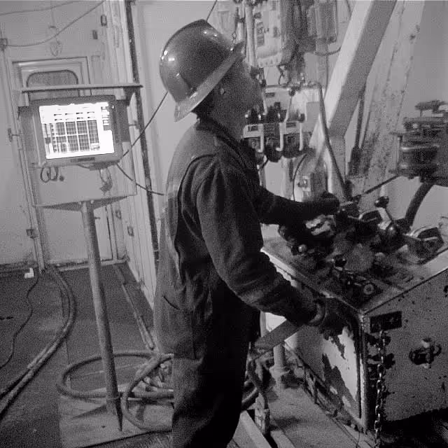 A worker wearing a hard hat and gloves operates machinery controls in an industrial setting.