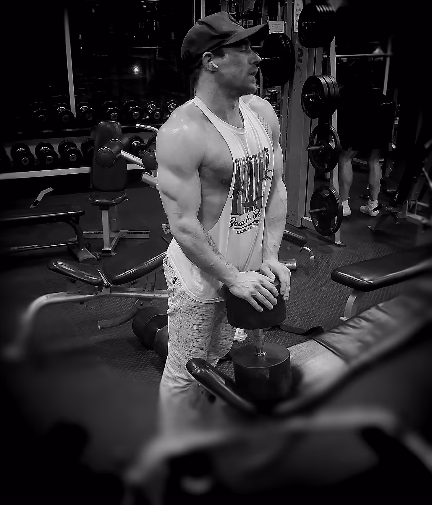 Alex, muscular  in a sleeveless shirt and cap holding a large dumbbell in the gym.