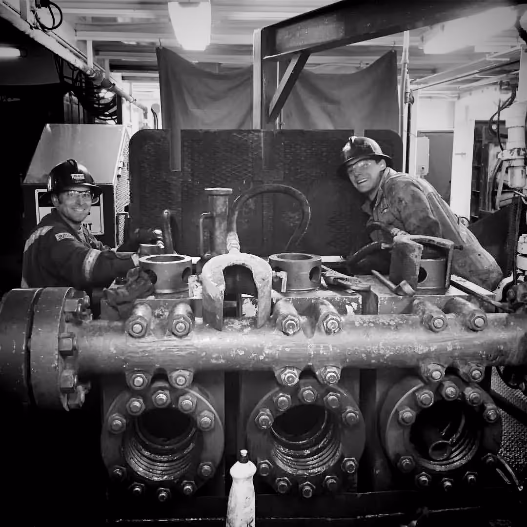 Two workers wearing hard hats and safety glasses smiling beside large industrial machinery inside a factory or plant.