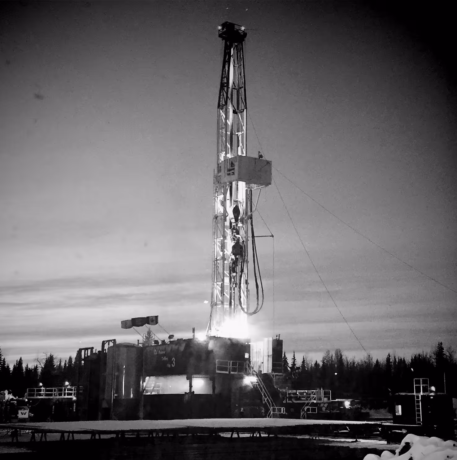 Illuminated oil drilling rig at dusk with trees and snow in the background.