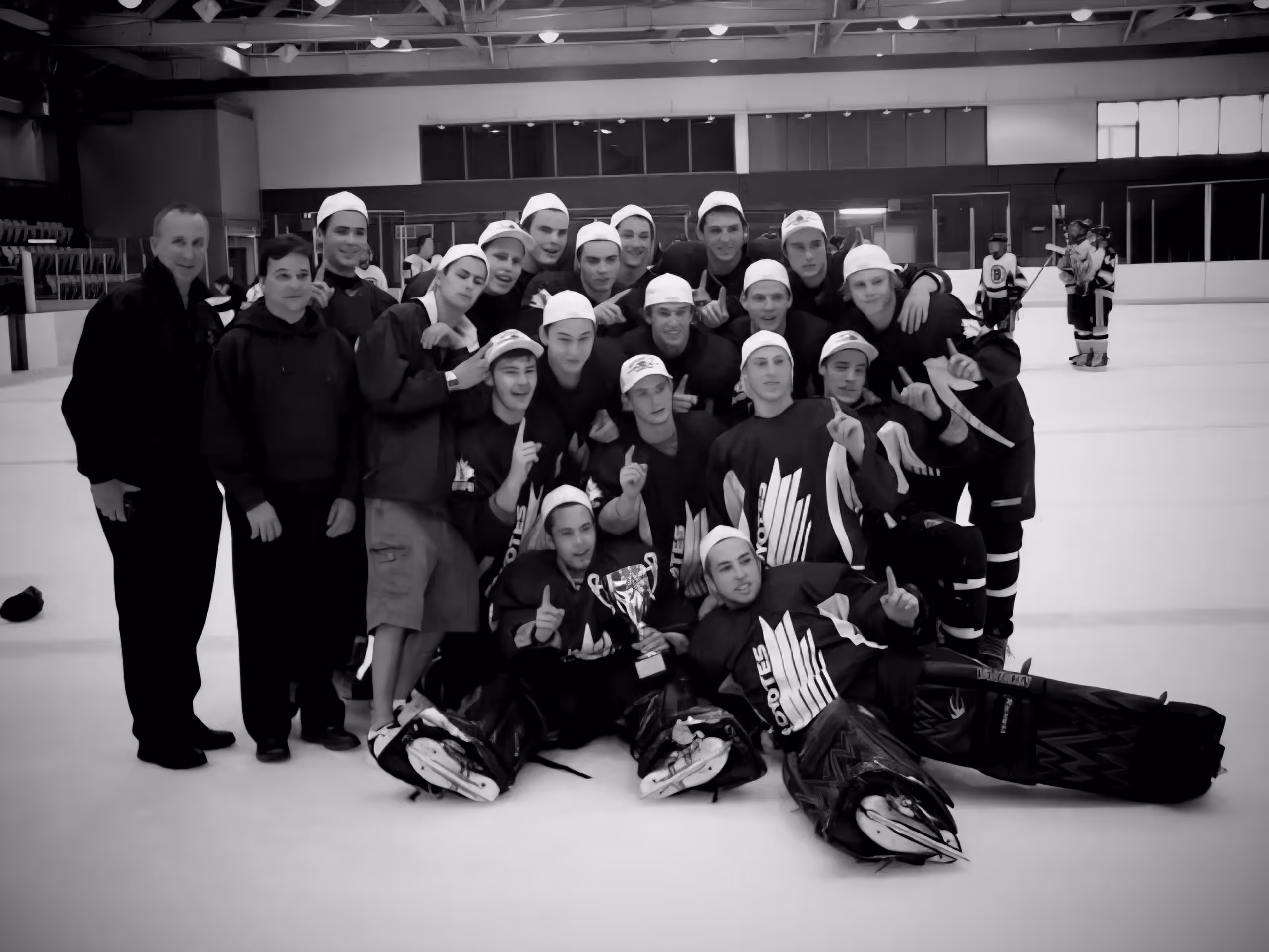 Ice hockey team posing on the ice rink with a trophy, wearing jerseys and caps, some players kneeling and holding up number one fingers.