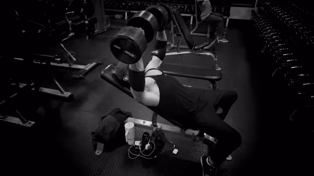 Alex in a gym performing seated dumbbell shoulder press with heavy weights.