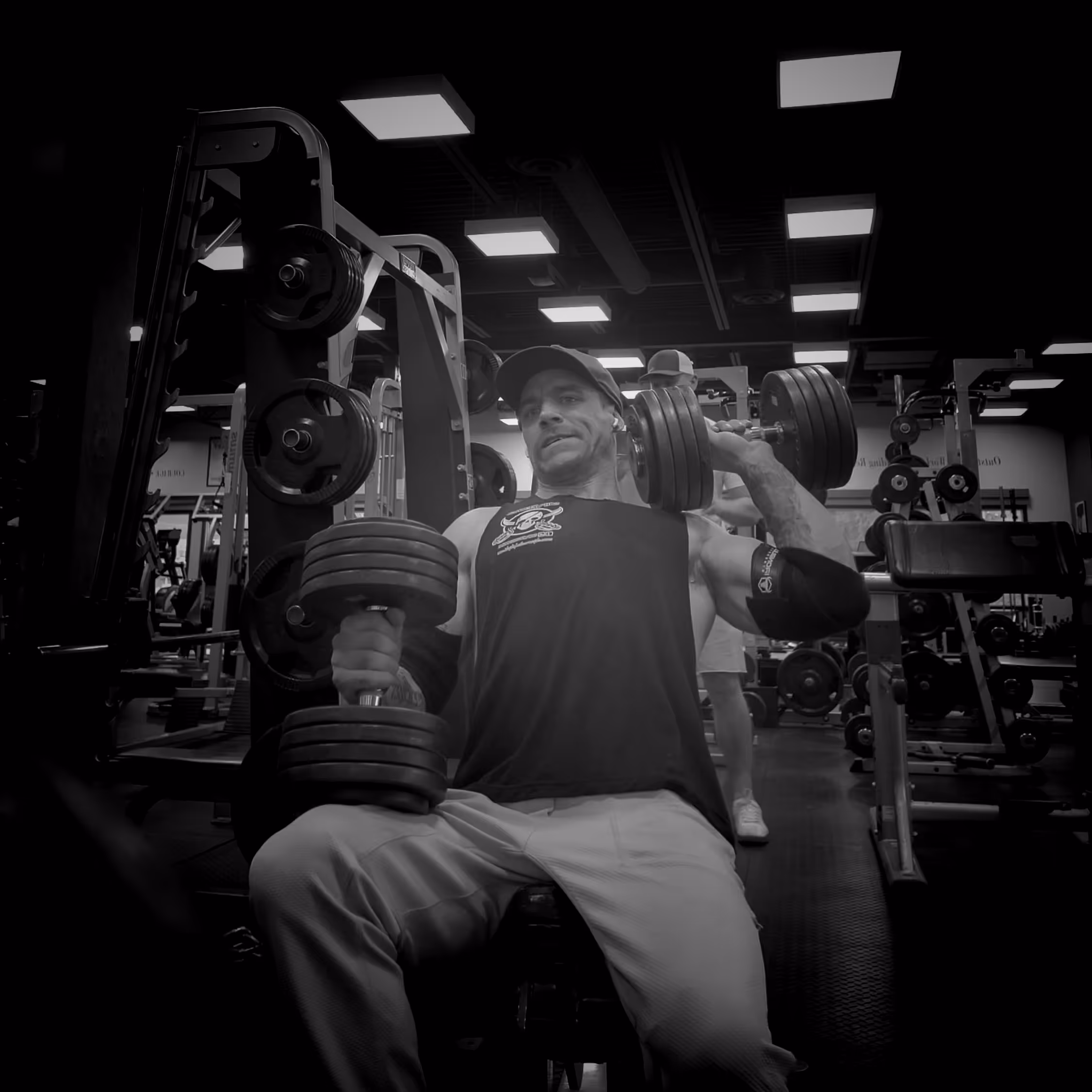 Man in gym wearing a tank top and cap lifting heavy dumbbells while seated, with weight machines in the background.