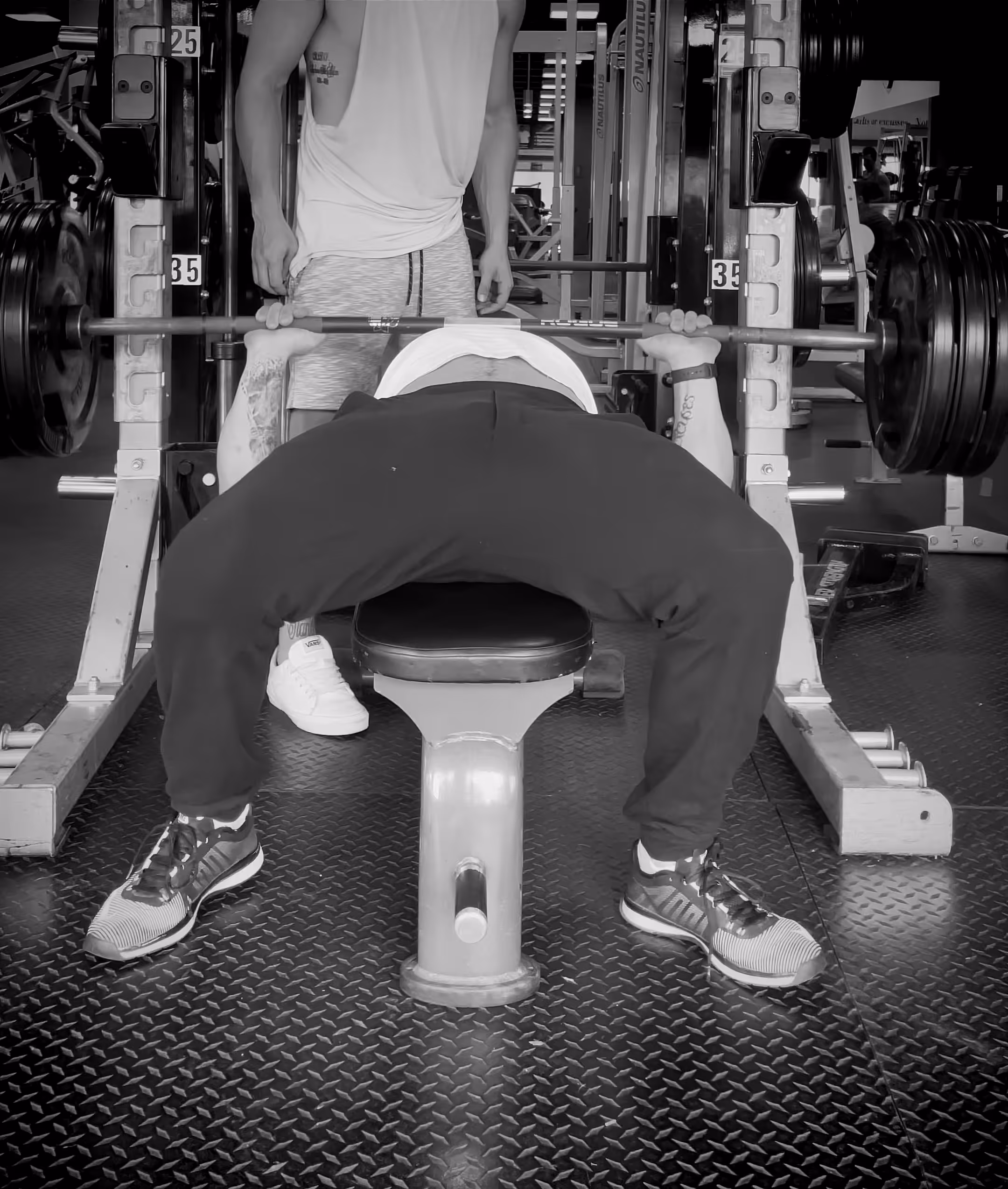Alex bench pressing heavy barbell with spotter standing behind in a gym.