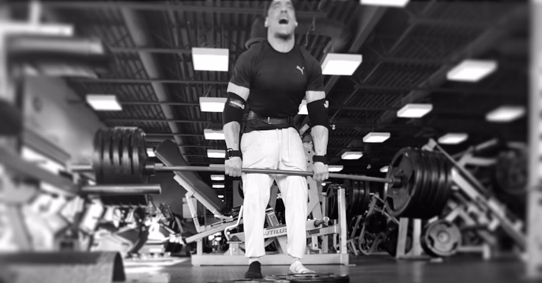Man in gym lifting a heavily loaded barbell while wearing weightlifting gear and showing intense effort.