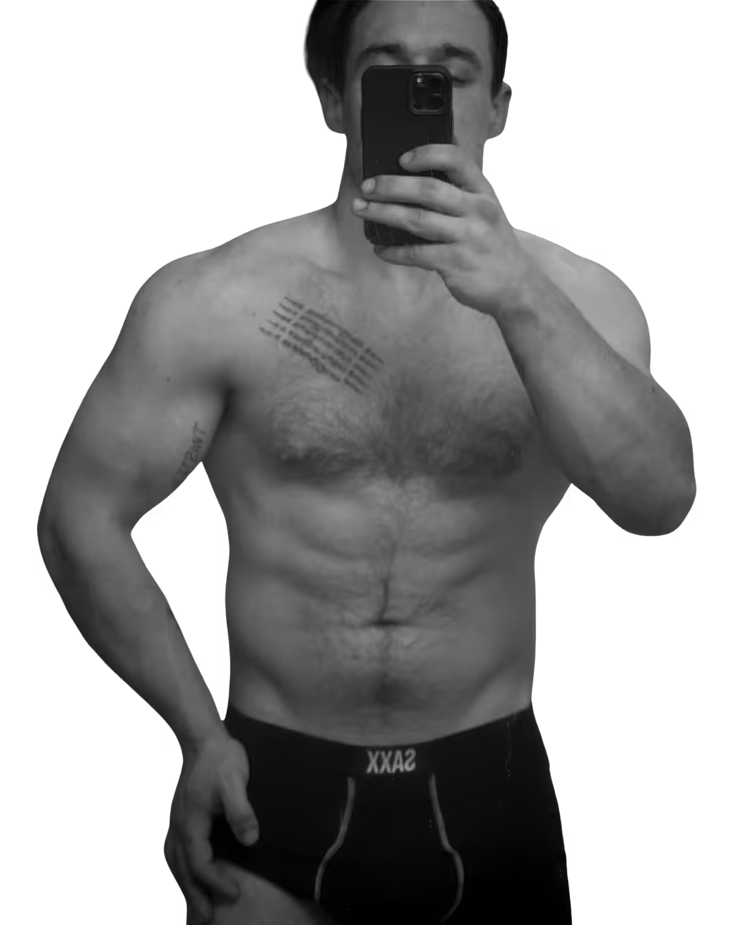 Shirtless muscular man taking a selfie with a smartphone, showing tattoos on his chest and arm, wearing black SAXX underwear.