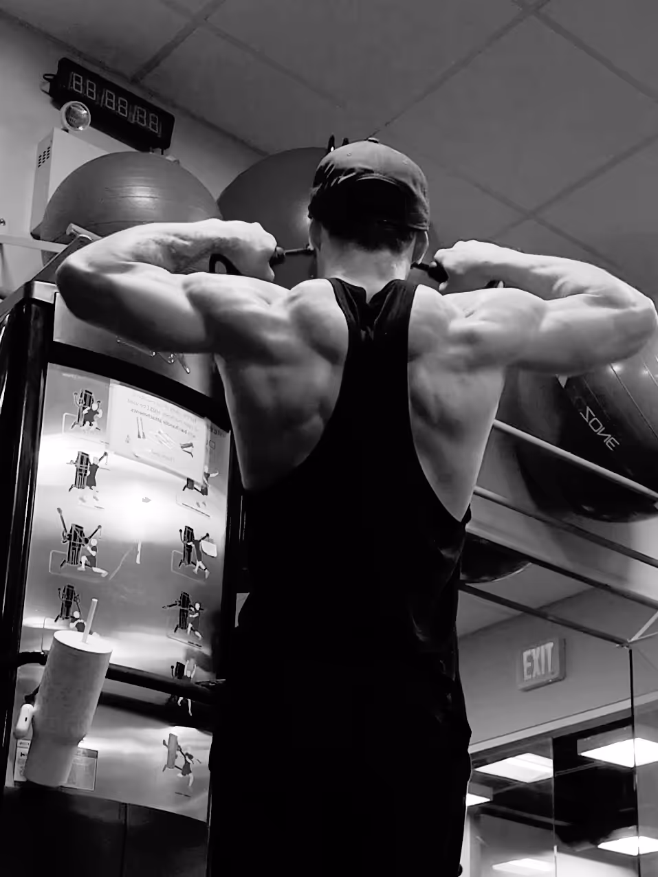 Man wearing a tank top and cap doing a pull-up exercise in a gym with fitness balls and mirrors in the background.