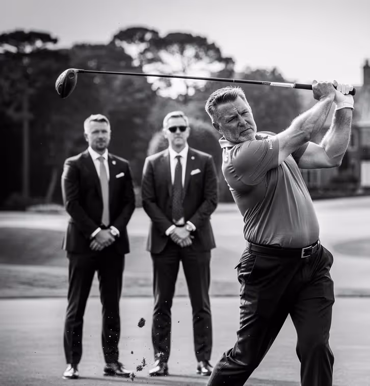 Man in golf attire swinging a golf club with two men in suits standing and watching in the background on a golf course.