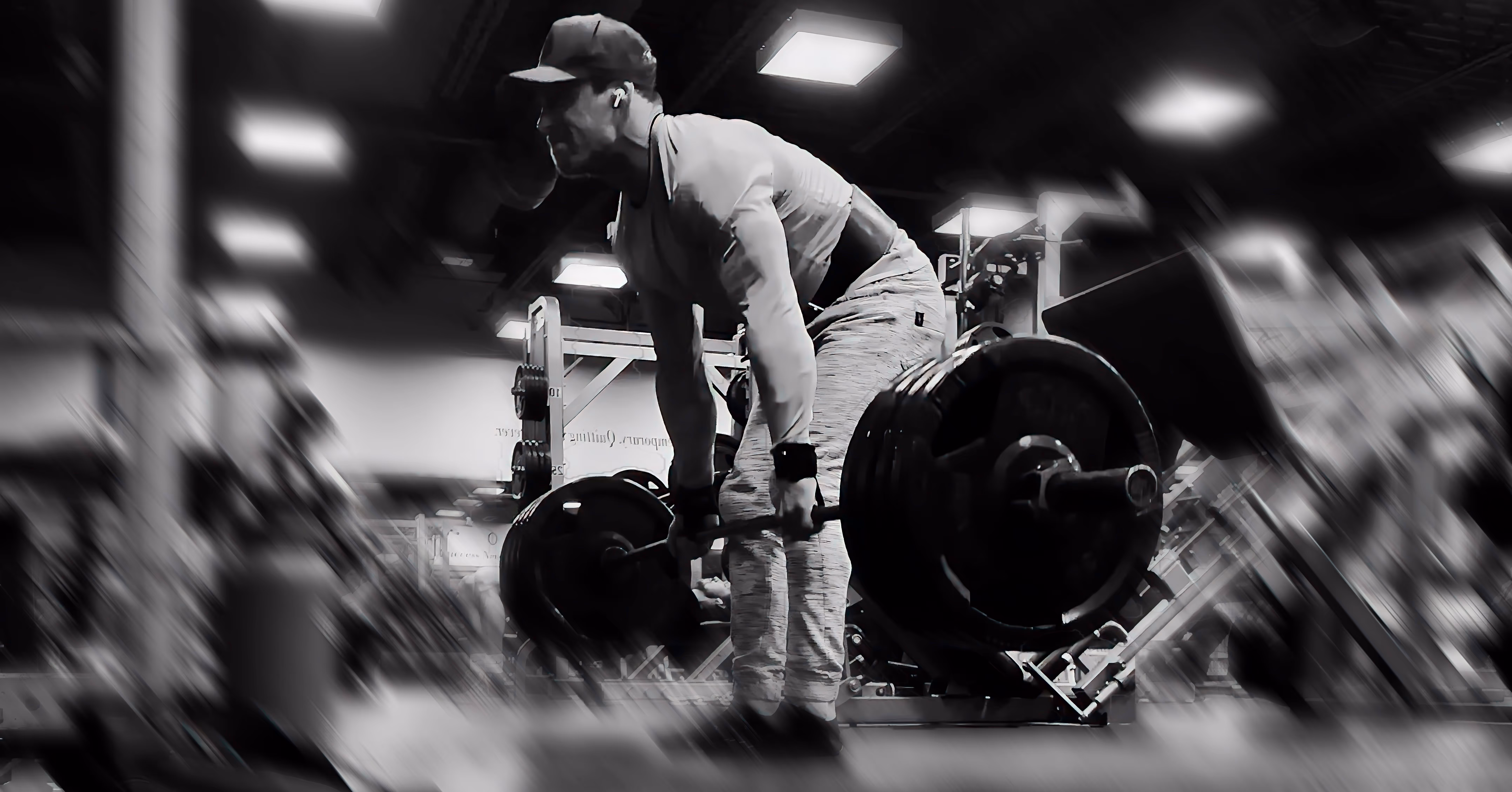 Alex wearing a cap and earbuds deadlifting a heavy barbell in a gym.