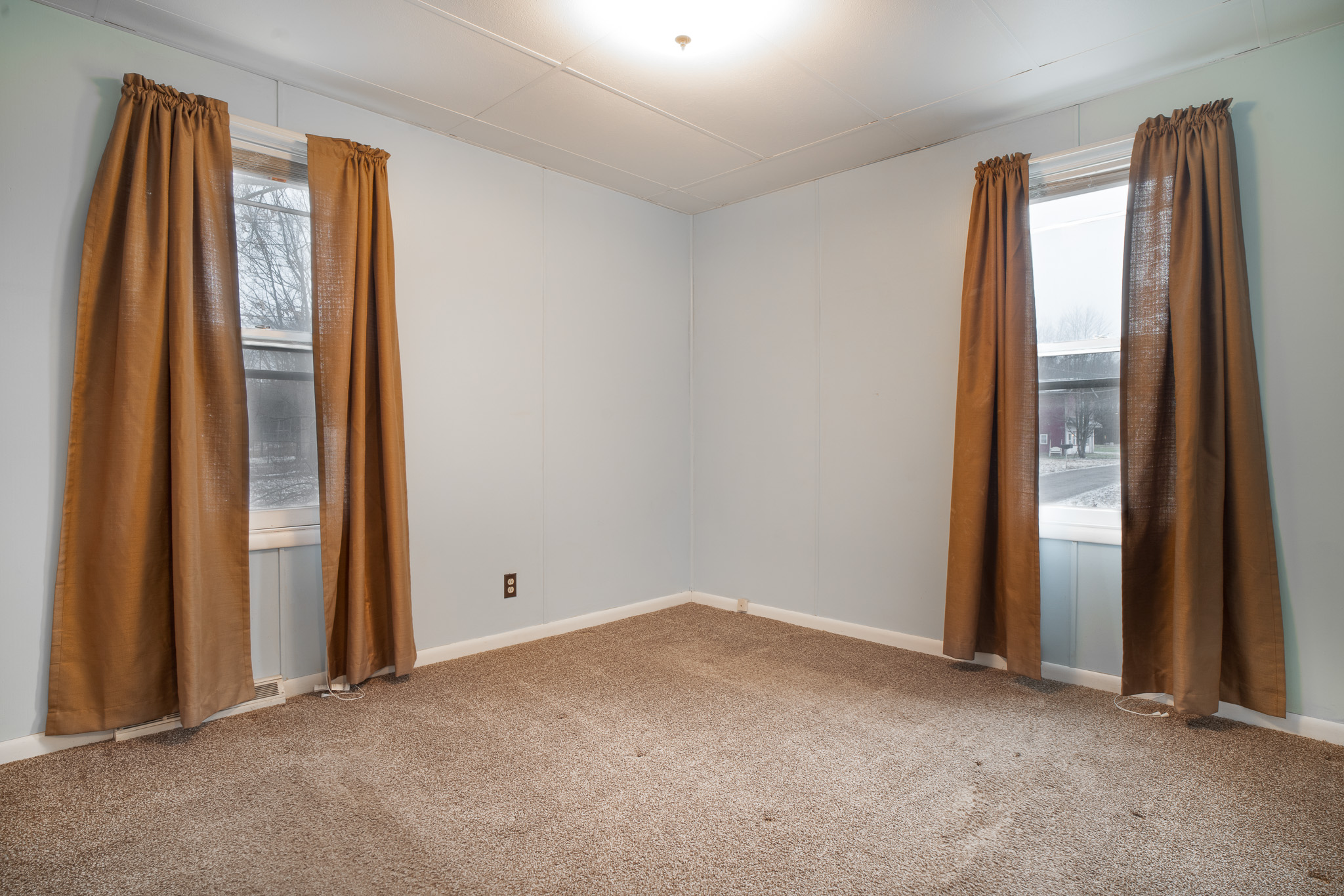 Empty corner room with light blue walls, brown carpet, and two windows with brown curtains.