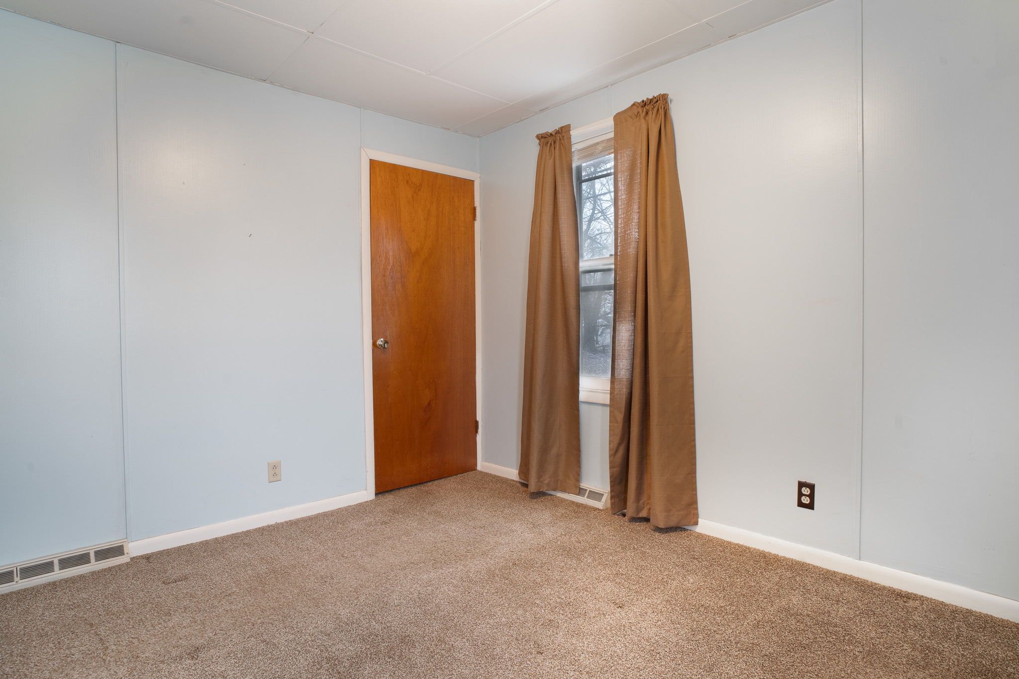 Empty room with beige carpet, light blue walls, a wooden door, and a window with brown curtains.