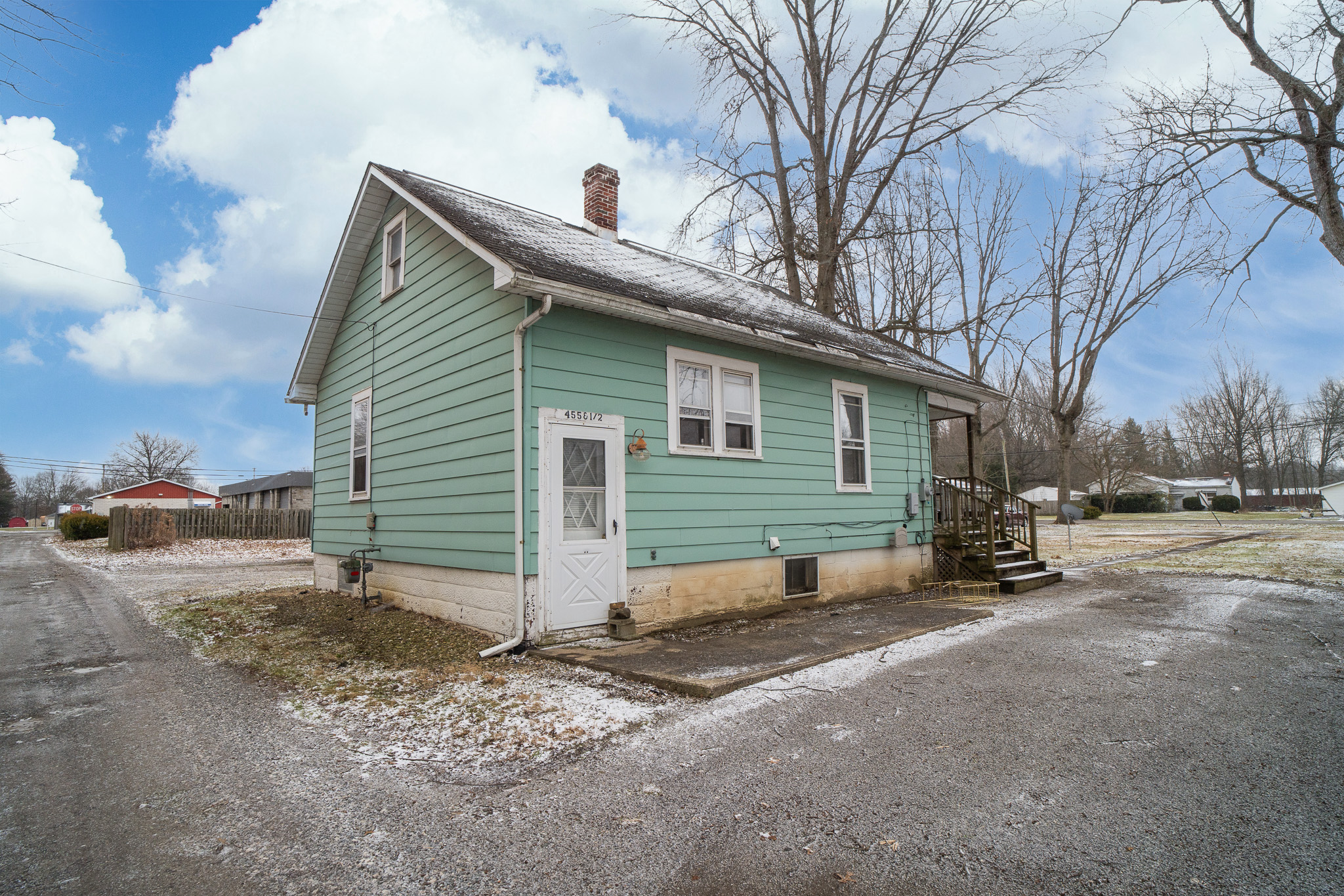 Small green house with white door and windows, light snow on roof and ground, leafless trees around.