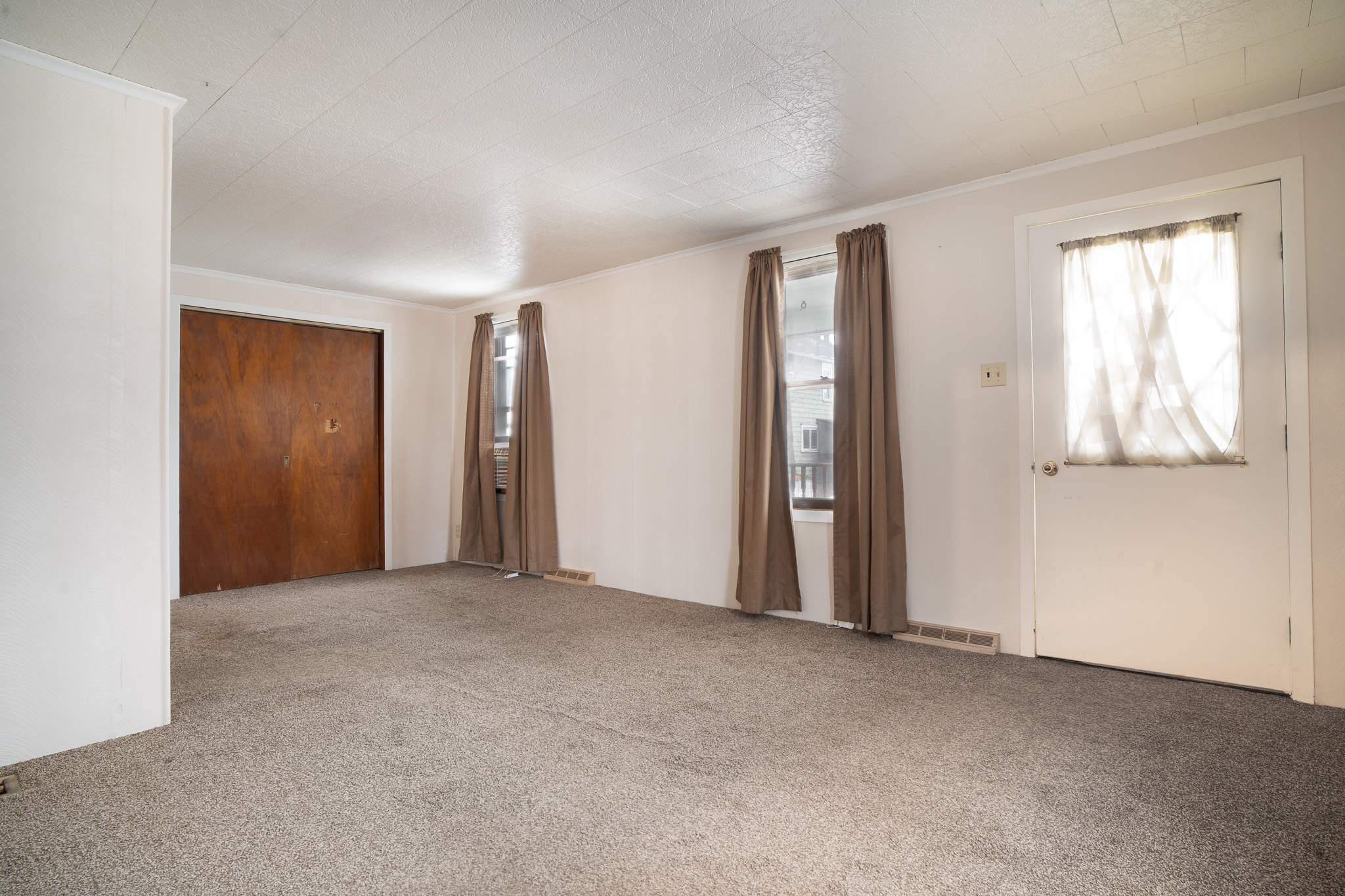 Empty room with beige carpet, white walls, two windows with brown curtains, a brown closet door, and a white door with a sheer curtain.