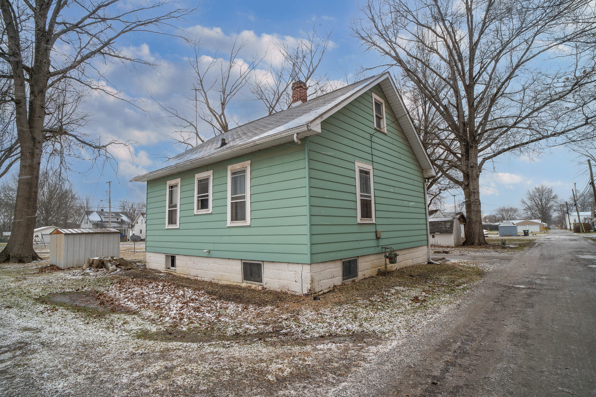 Small green house with white trim on a snowy yard beside a dirt road with leafless trees.