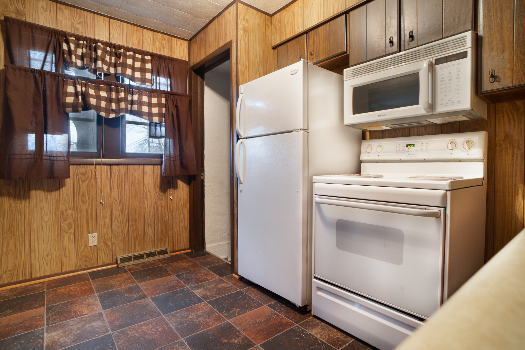 Kitchen corner with white refrigerator, stove, microwave, wood-paneled walls, and tiled floor.