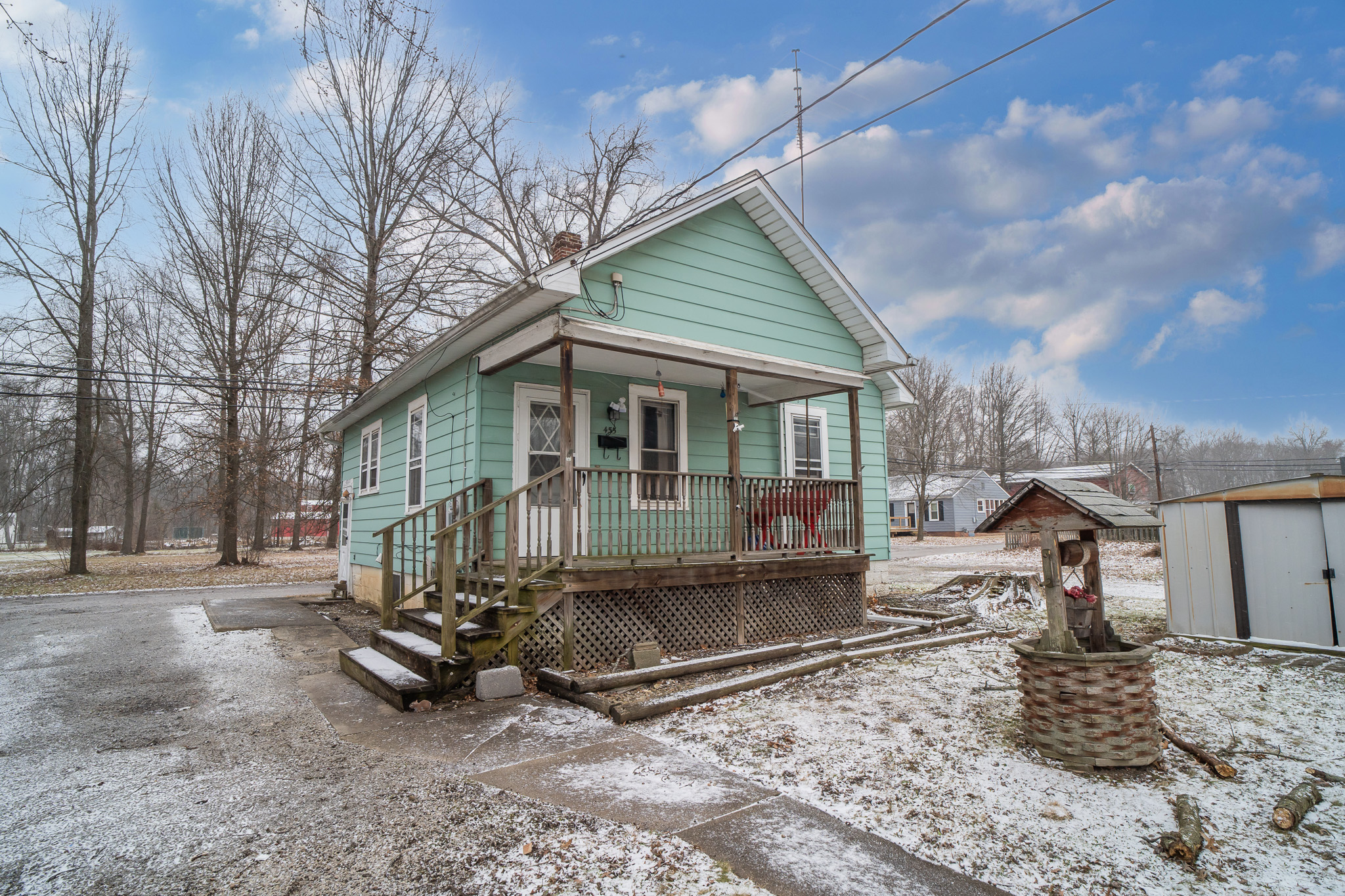 Small light green house with a wooden porch and steps, bare trees, light snow on the ground, and a decorative wooden well in the yard.