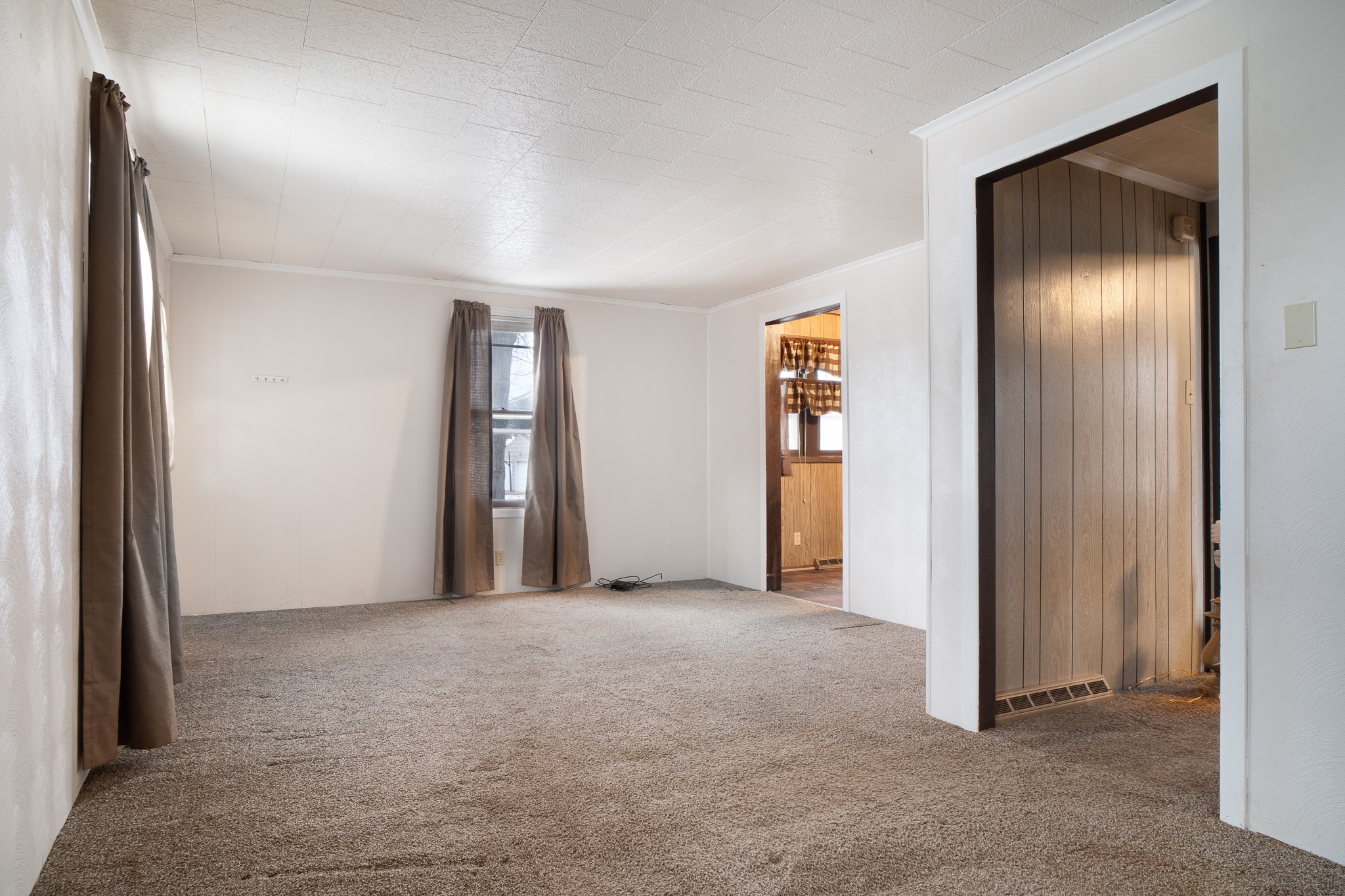 Empty room with beige carpet, white walls, two small windows with brown curtains, and doorways leading to other rooms.