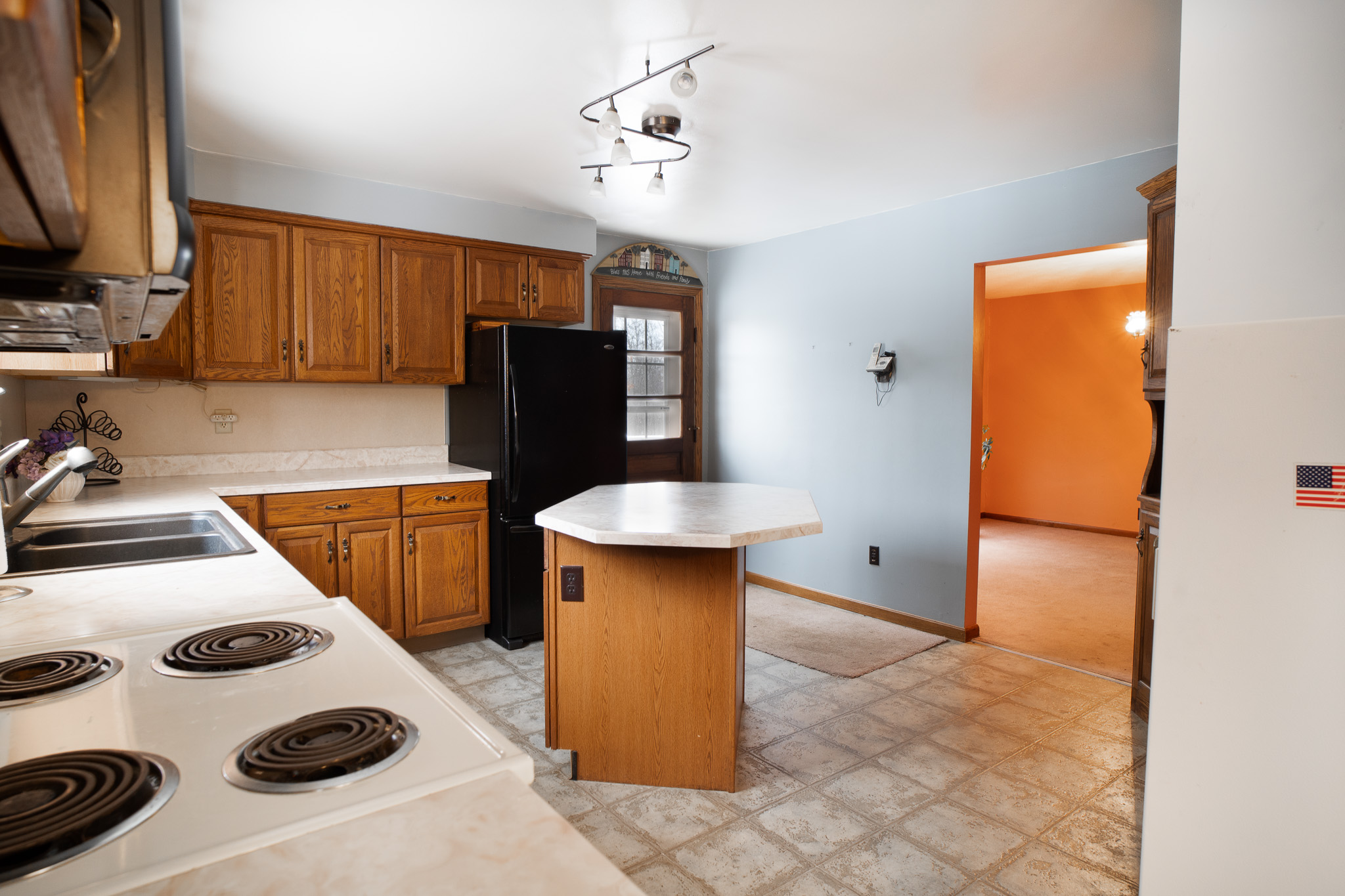 Kitchen with wooden cabinets, a black refrigerator, an octagonal island with a marble countertop, and a white electric stove, adjacent to a room with orange walls.