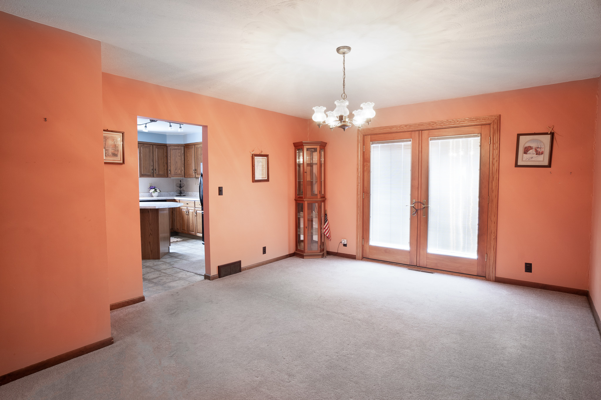 Empty living room with peach-colored walls, beige carpet, a chandelier, and French doors with blinds leading outside, adjacent to a kitchen with wooden cabinets.