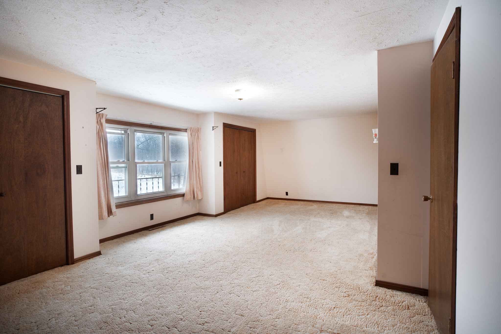 Empty beige carpeted room with three brown wooden doors, a large window with light curtains, and a ceiling light fixture.