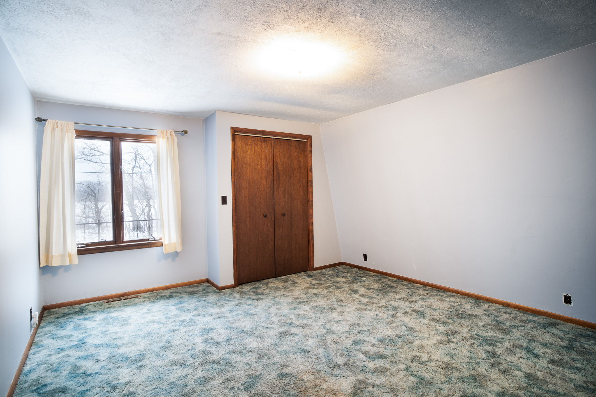 Empty room with blue-green carpet, white walls, a window with cream curtains, and a wooden double-door closet.