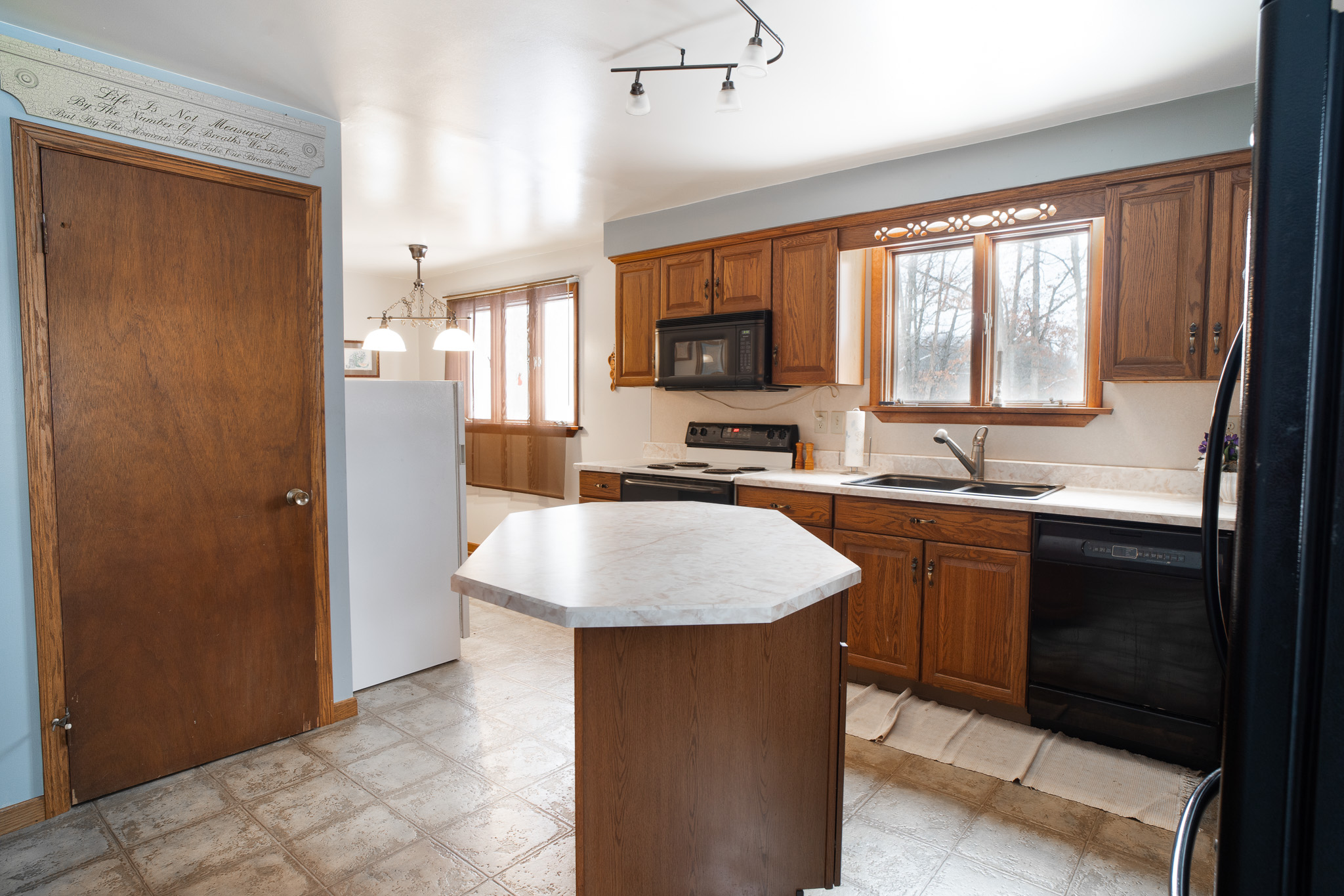 Bright kitchen with wooden cabinets, white countertops, central island, black appliances, and a window above the sink.