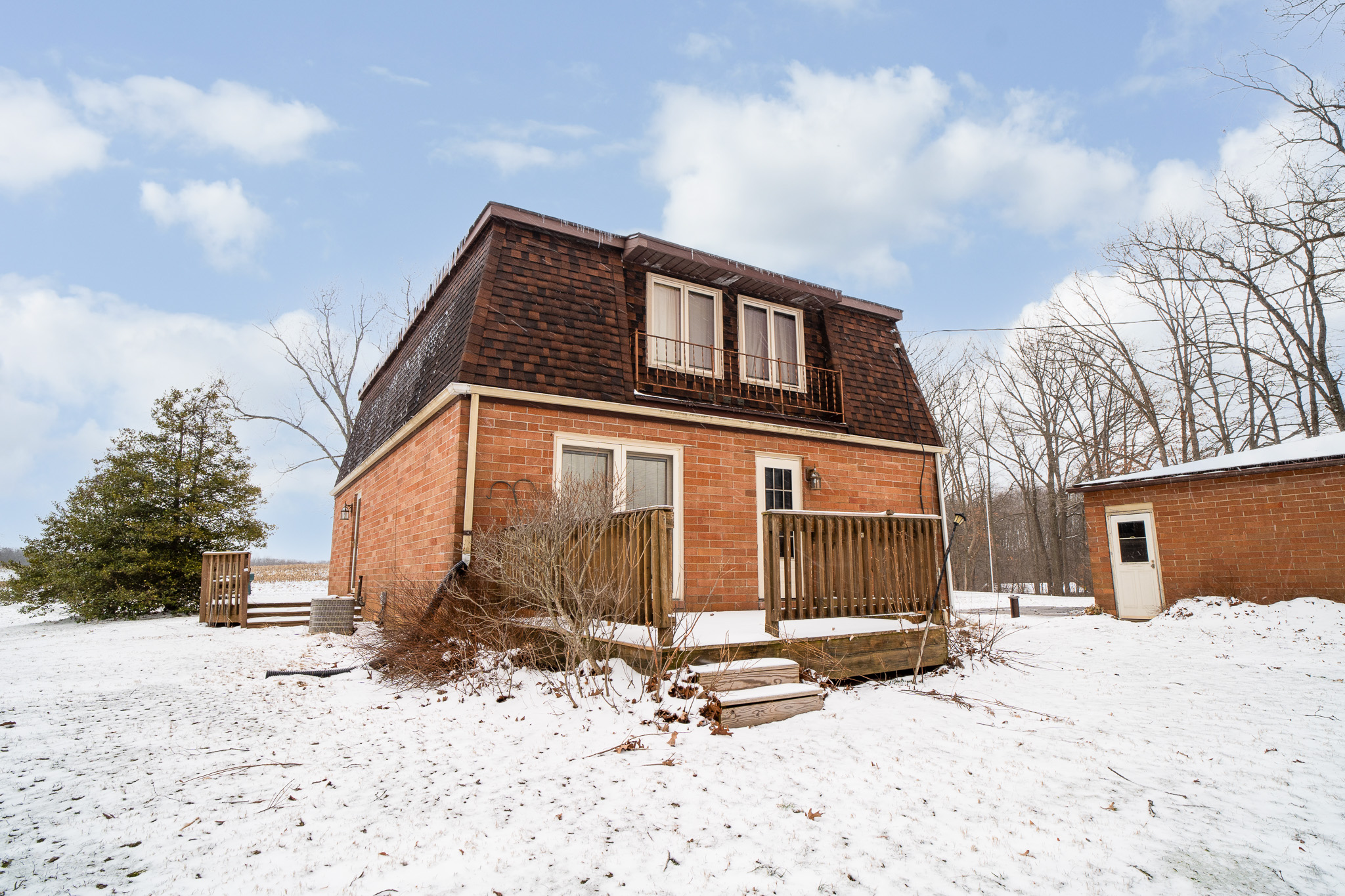 Two-story brick house with dark shingled mansard roof and wooden porch in a snow-covered yard under a cloudy blue sky.