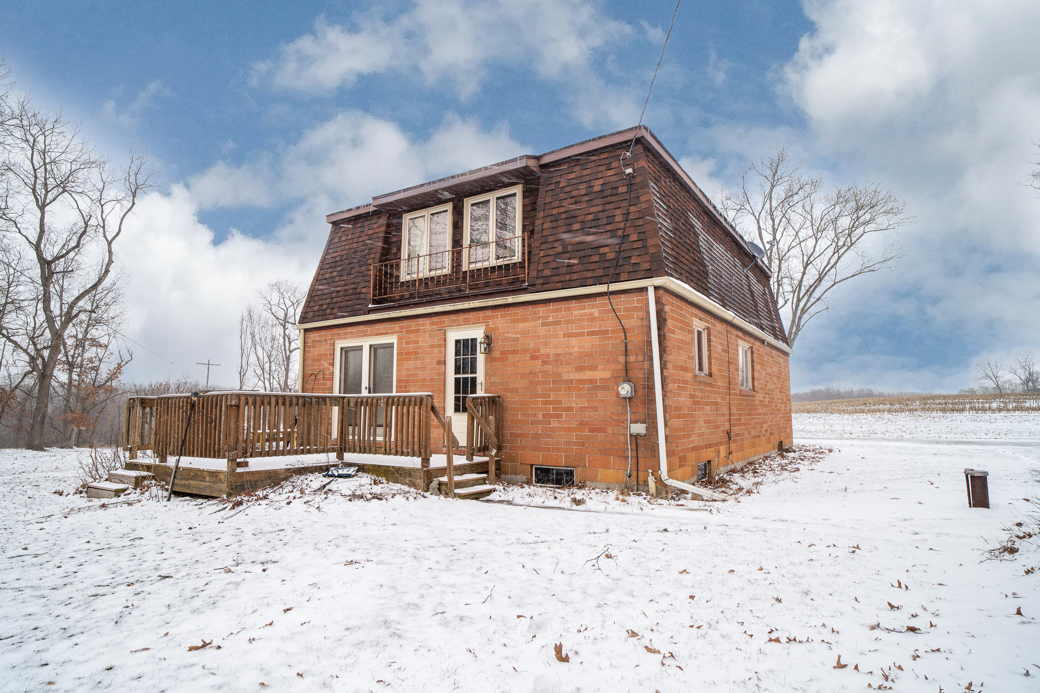 Brick house with a brown shingled mansard roof and wooden deck surrounded by snow on a cloudy winter day.