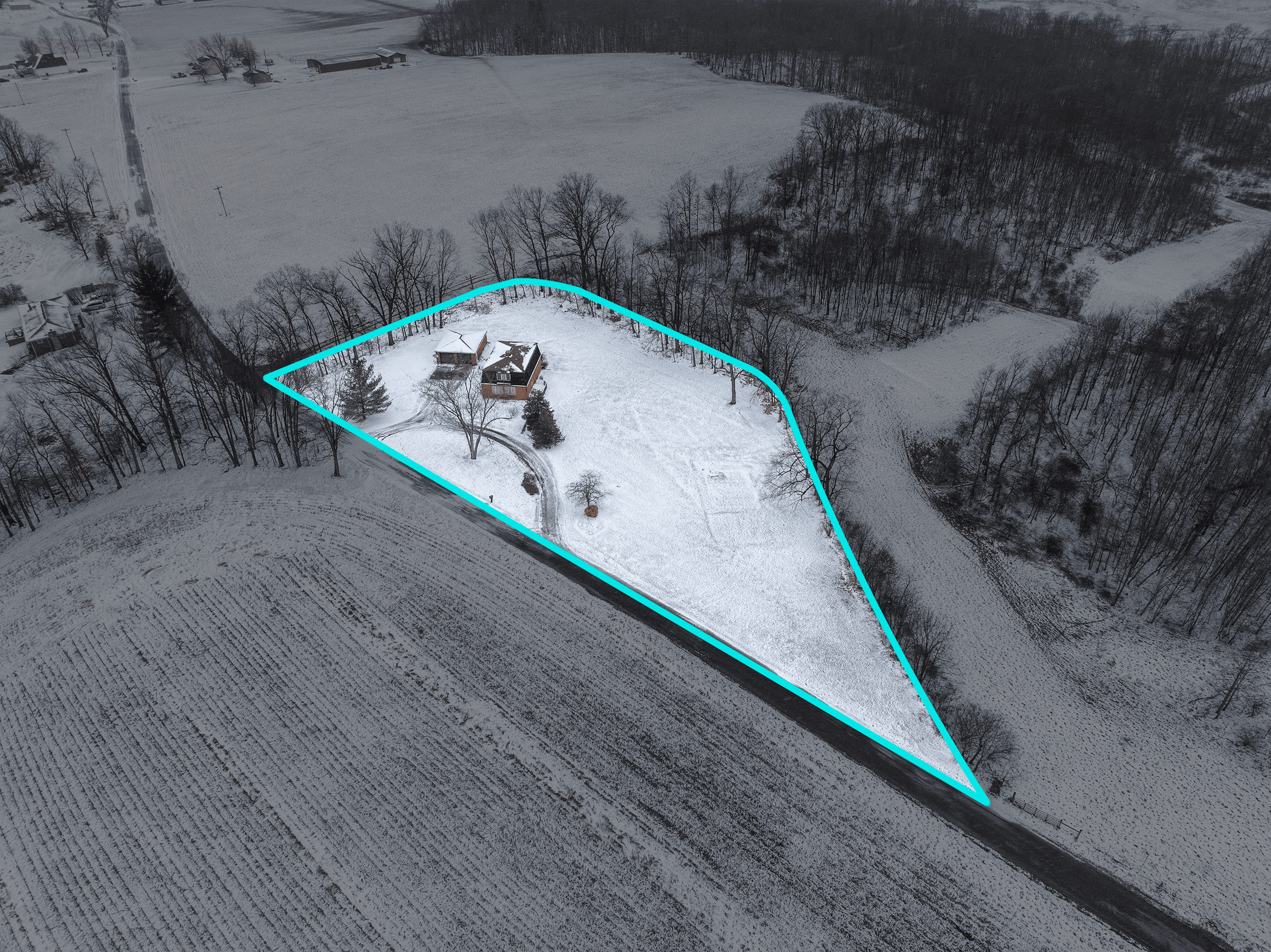 Aerial view of a snow-covered triangular property outlined in bright blue, featuring two houses, trees, and a curved driveway, surrounded by fields and wooded areas.