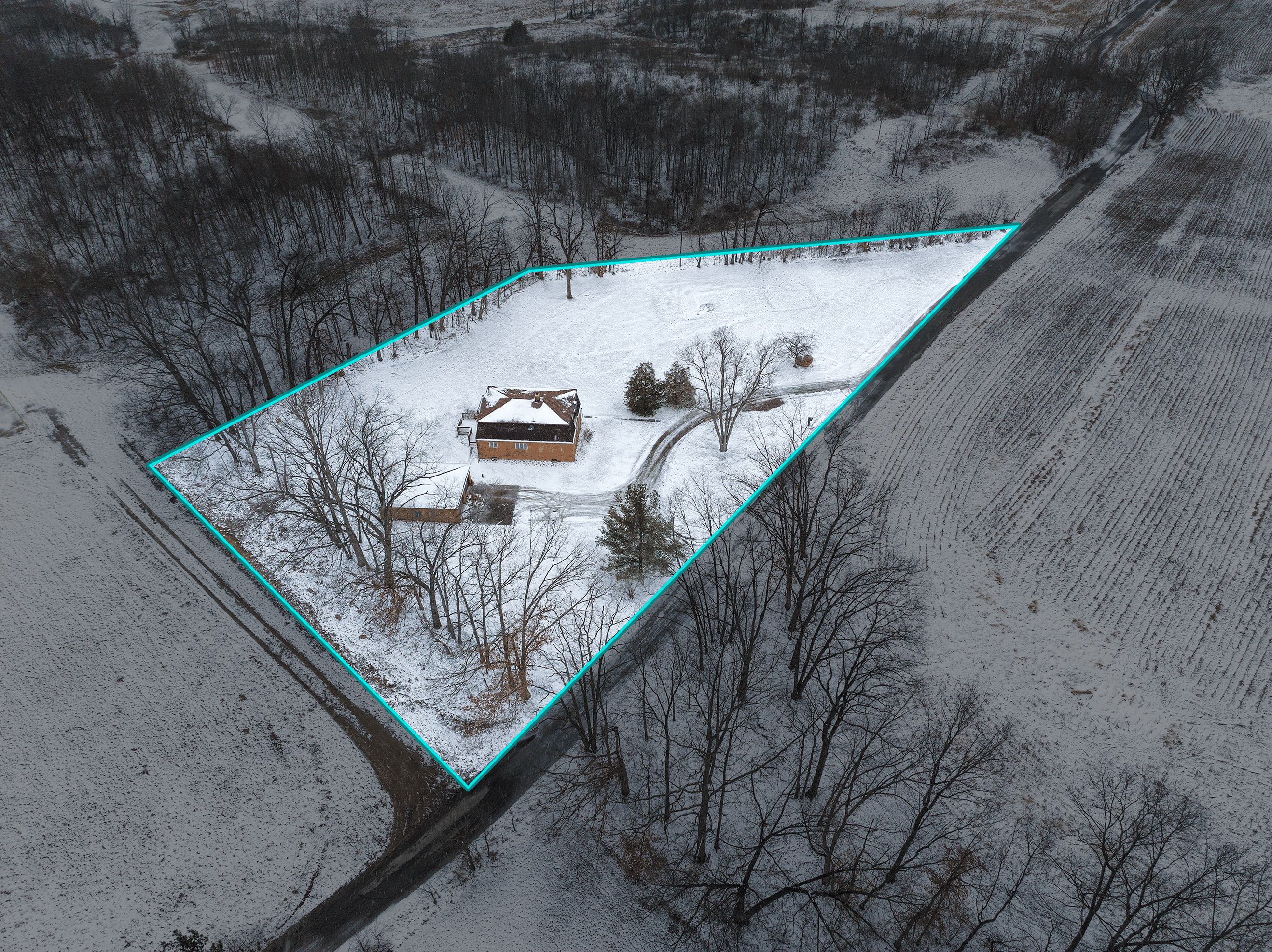 Aerial view of a snow-covered house and yard outlined by a turquoise border surrounded by leafless trees and fields.