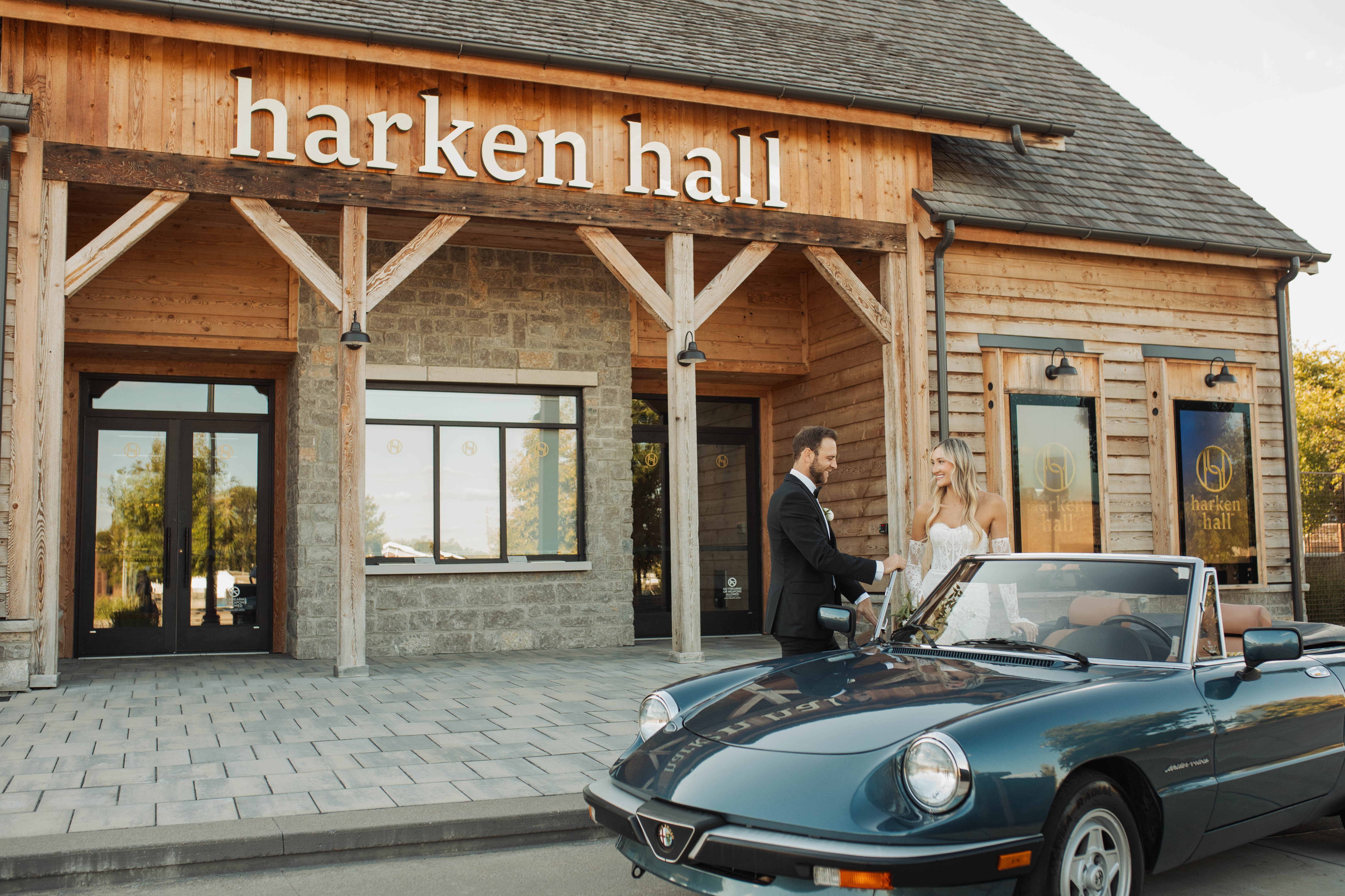 An expansive outdoor patio and courtyard at Harken Hall, featuring long wooden bar counters, rows of black dining tables and chairs, string lights overhead, and a view of the modern venue architecture surrounded by green trees.