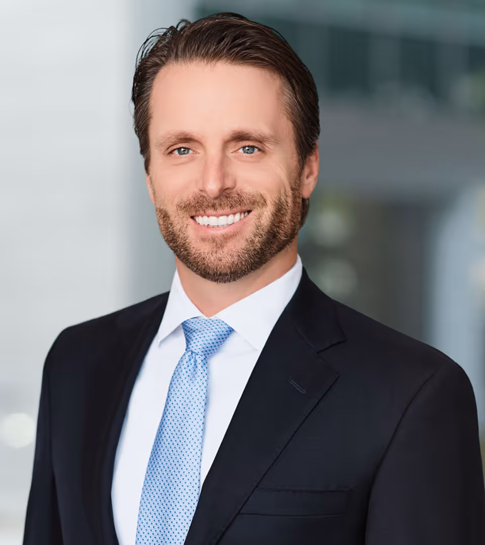 Smiling man with light beard wearing a dark suit, white shirt, and light blue patterned tie against a blurred background.
