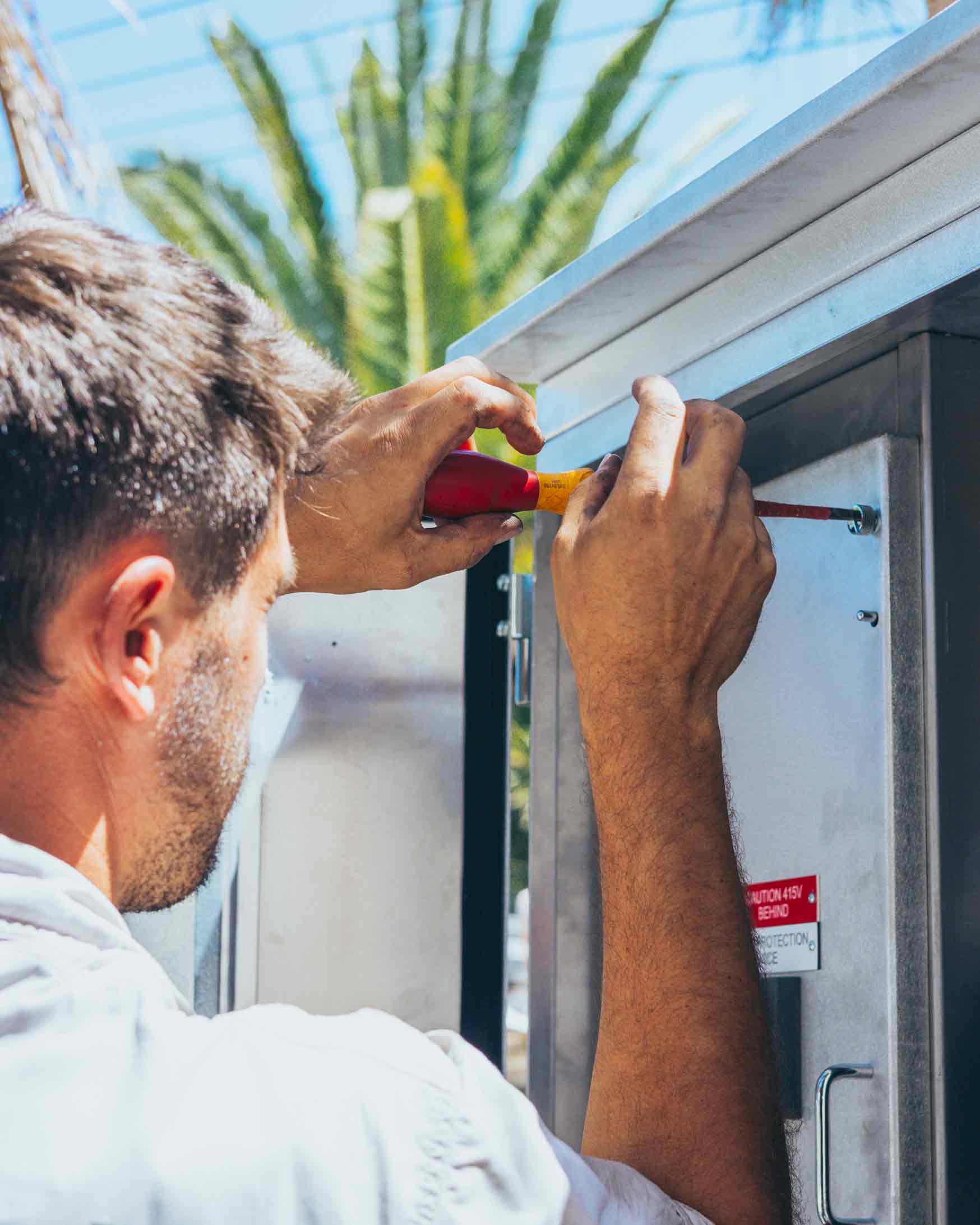 Electrical technician using a screwdriver to repair an outdoor electrical switchboard mounted on a white wall.