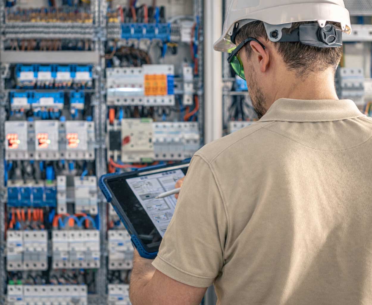 Electrical technician using a screwdriver to work on an open outdoor electrical switchboard mounted on a white brick wall.