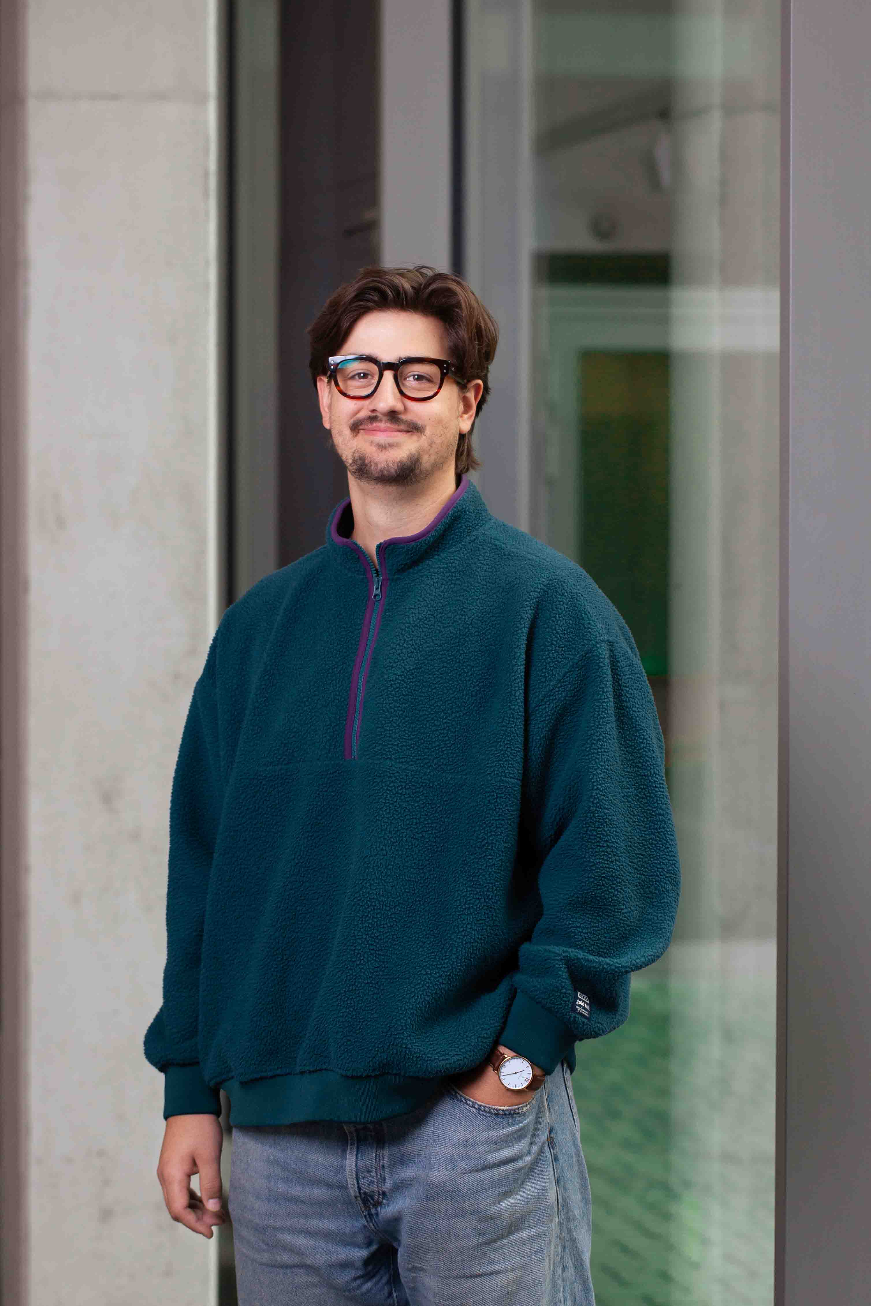 A yound man in jeans and pullover is standing in front of a window, looking into the camera and smiling