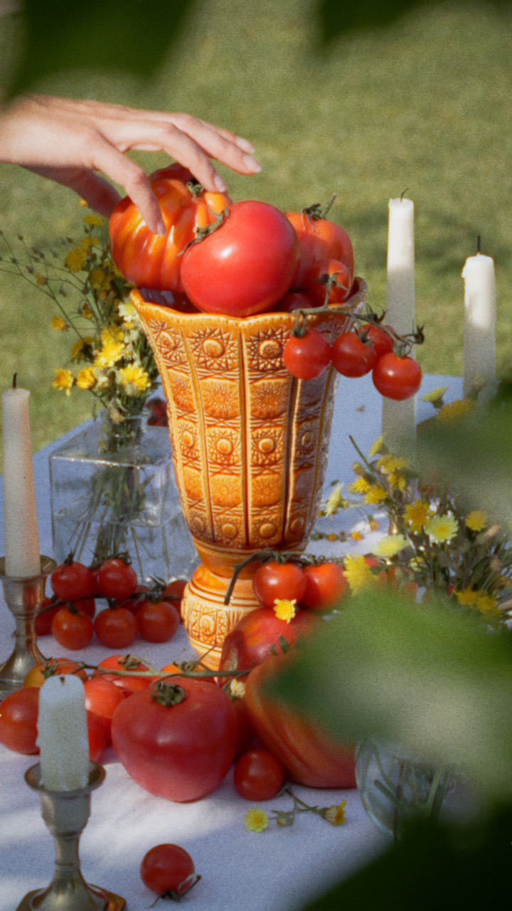 A ceramic jar on a table in a garden. The jar is filled with tomatos